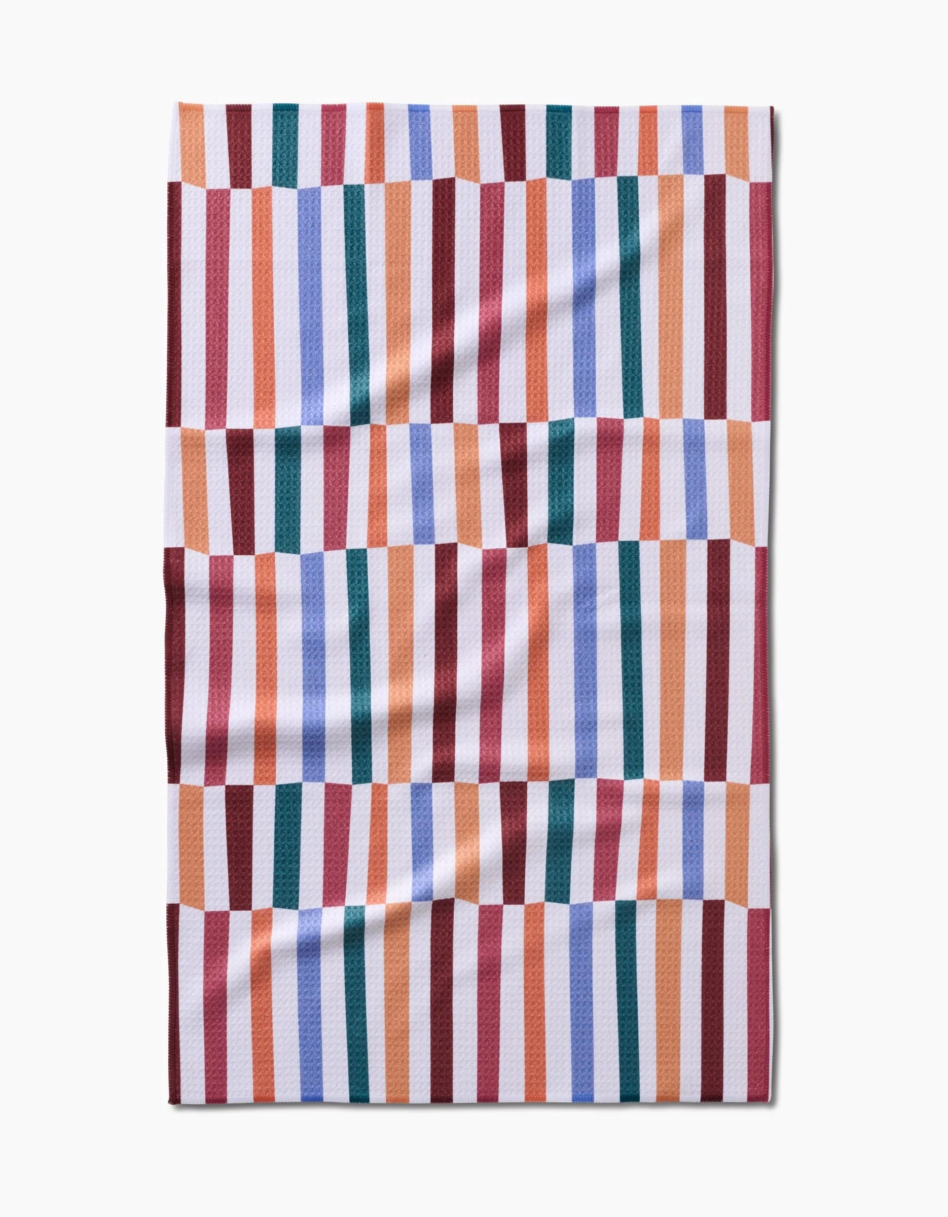 Claire Iglesias’ Wonky Stripes blanket, crafted from recycled materials, features bold vertical stripes in red, orange, blue, green, lavender, and white laid flat against a white background in a colorful geometric pattern.