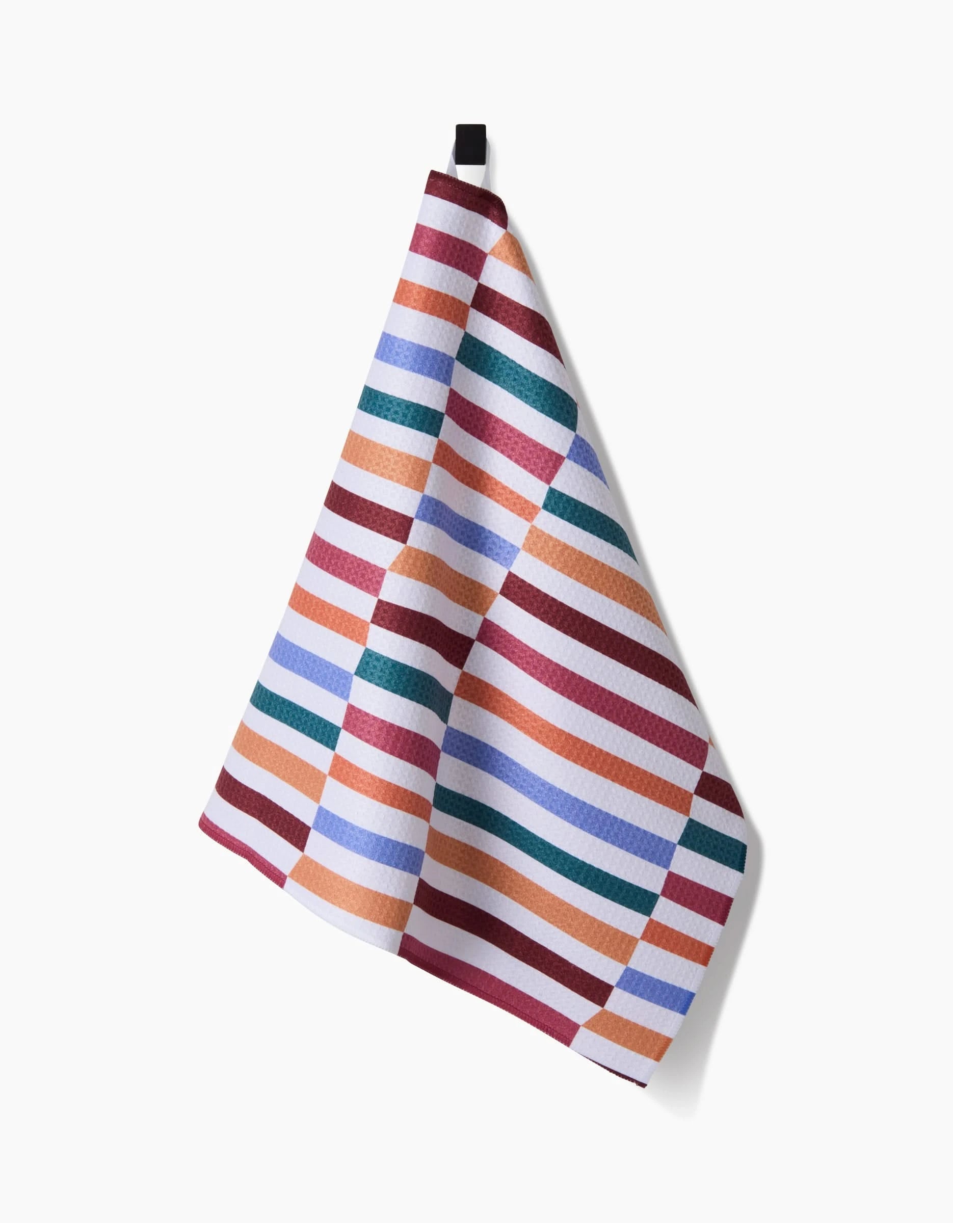 The Wonky Stripes tea towel by Claire Iglesias features horizontal red, orange, blue, purple, and teal stripes. Made from recycled materials, it hangs from a black loop against a white background.