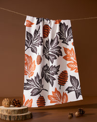The Geometry Woodland Waltz kitchen tea towel, made from recycled materials, showcases orange and black leaf and pinecone patterns. It hangs above a brown background with pinecones and wooden slices arranged below.