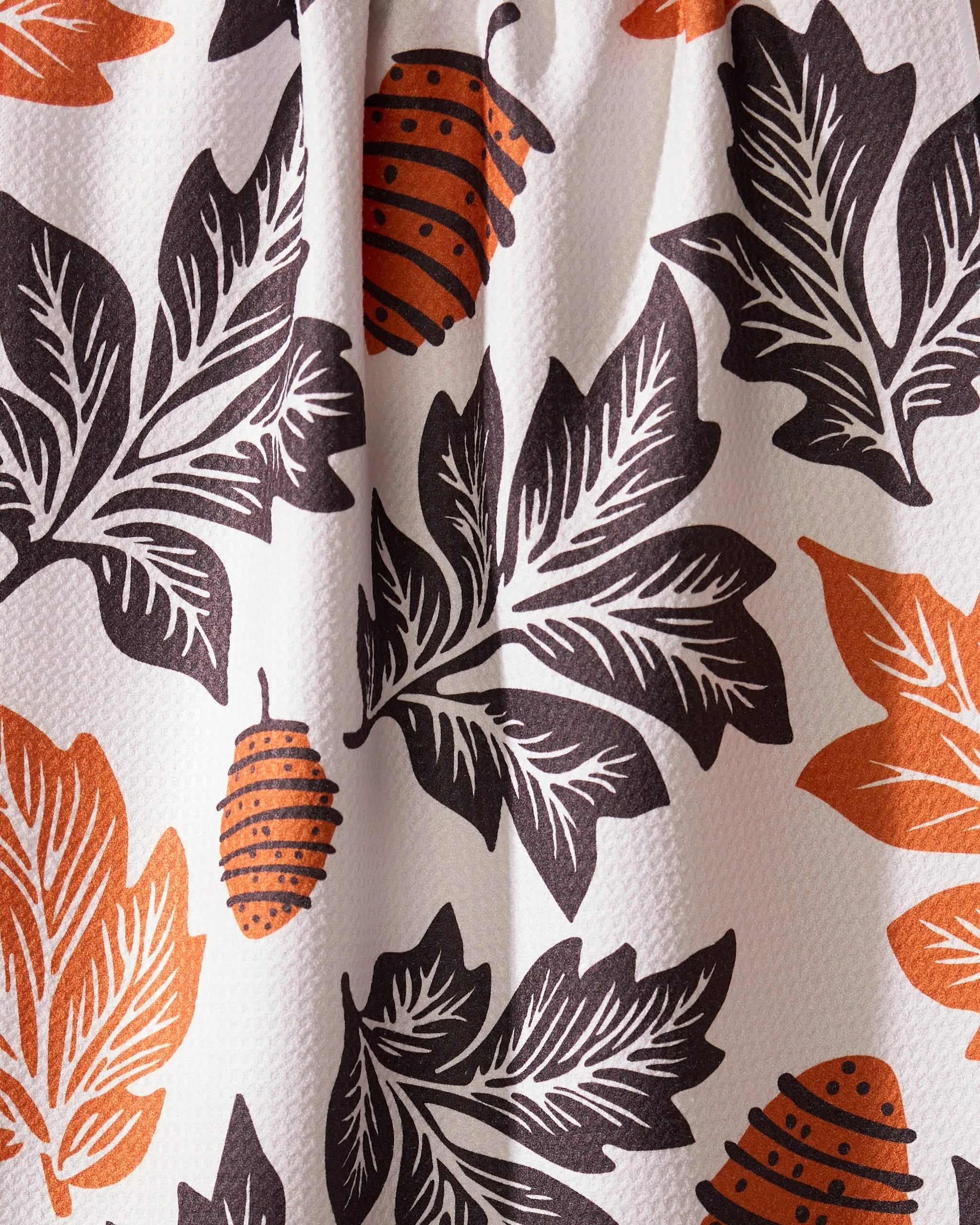 The Geometry Woodland Waltz tea towel features a white fabric with large black and orange leaves and small orange pine cones. It has a slightly bumpy texture and drapes softly with visible folds.