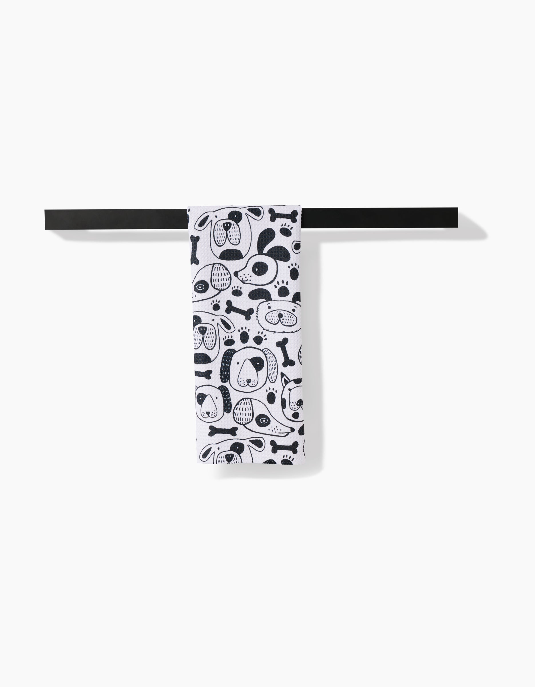 The Jessica Swift Woofin Around towel, featuring black cartoon dog faces and bones on white ultra-absorbent Geoweave fabric, hangs neatly on a black towel bar against a white background.