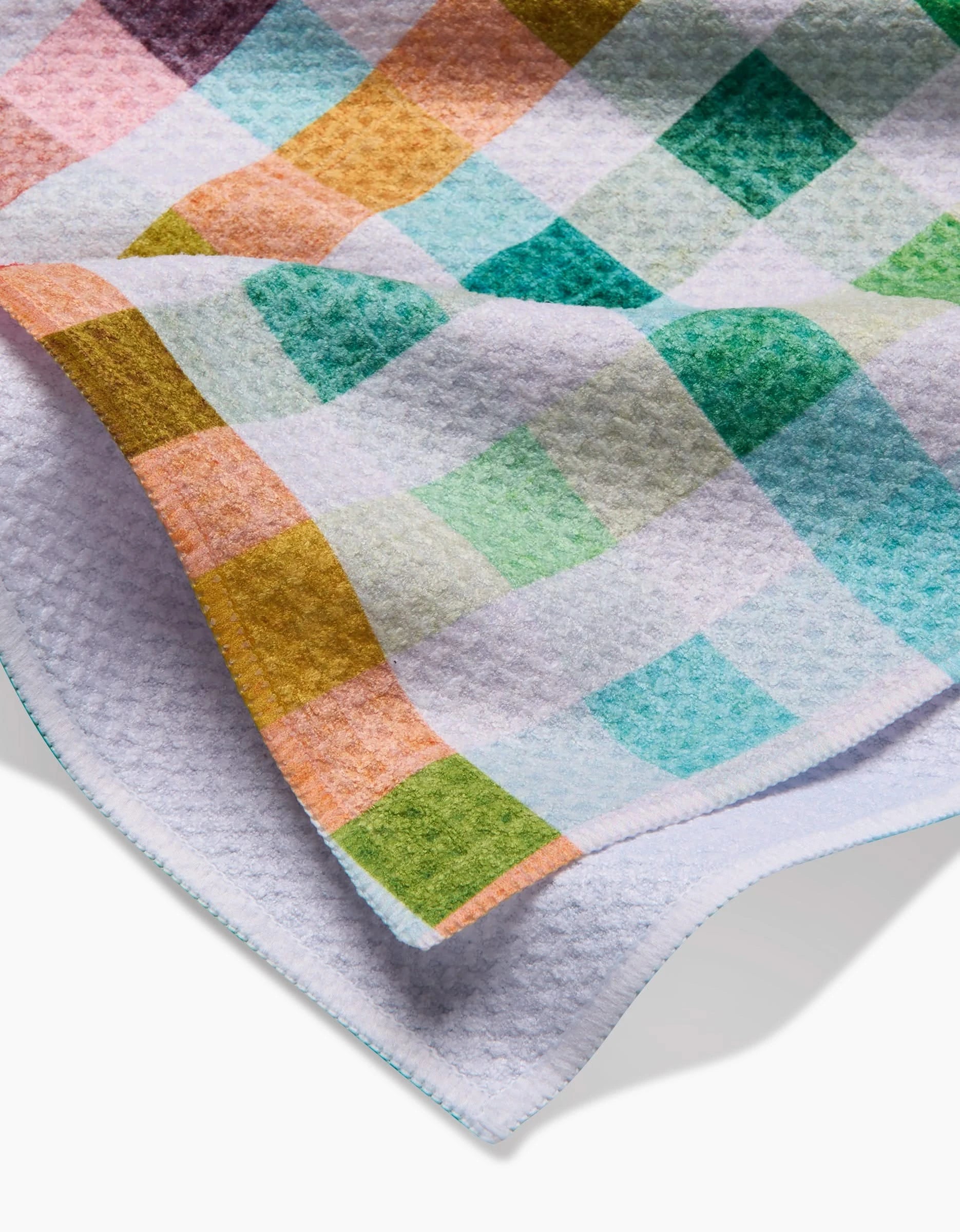 A close-up of the Pascale Dilger Woven Spring Plaid, a soft, ultra-absorbent blanket made from eco-friendly Geoweave fabric, features a colorful green, blue, yellow, and pink checkered pattern and is slightly folded on a white surface.
