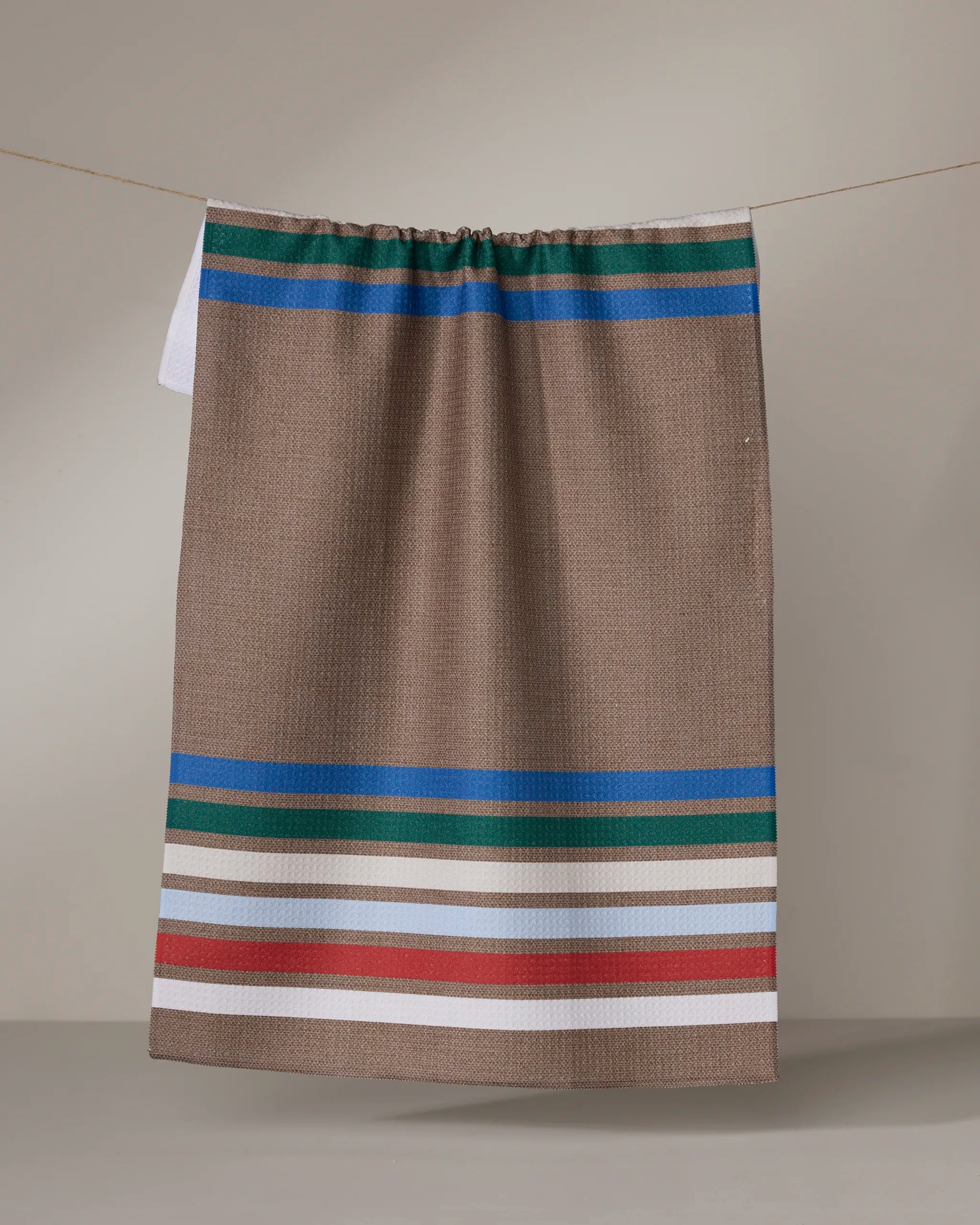 The Geometry Yuletide Band kitchen tea towel, featuring beige fabric with green, blue, white, and red horizontal stripes, hangs on a line against a neutral background.