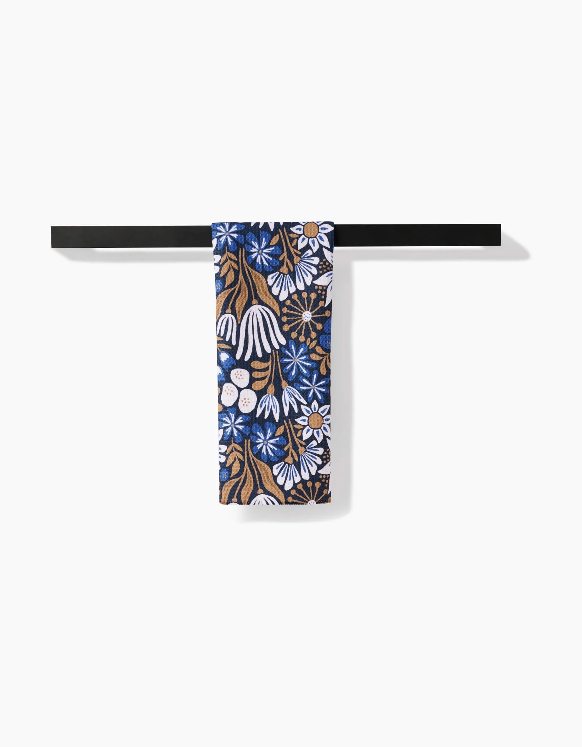 The Zillia tea towel by Amy MacCready features a blue, white, and brown floral design on soft Geoweave fabric for extra absorbency, and is shown hanging on a black towel bar against a white background.