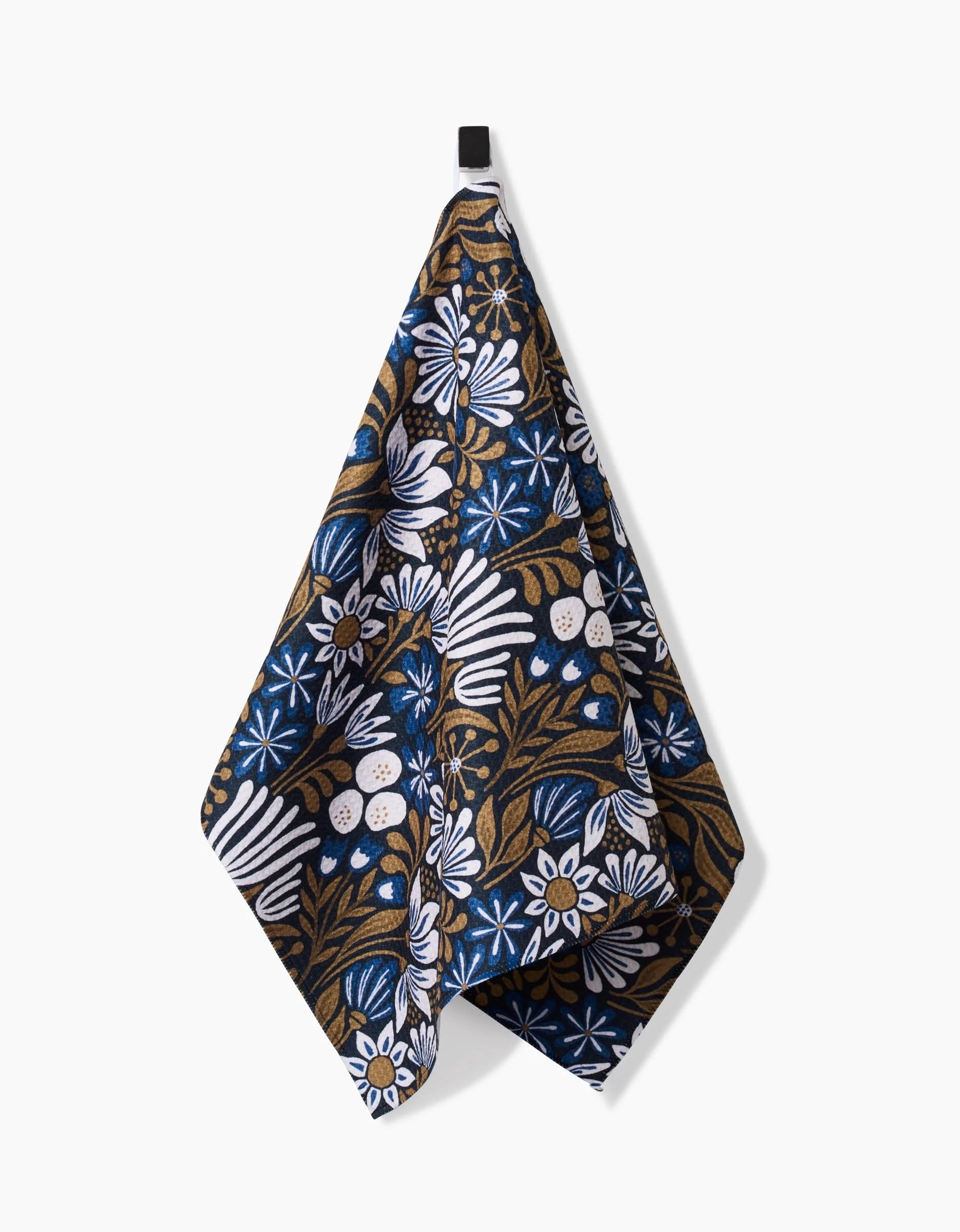 The Zillia Tea Towel by Amy MacCready hangs from a black hook. Made with Geoweave fabric, it features white flowers and green leaves on a dark blue background.