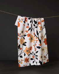 The Geometry Spiced Florals tea towel, featuring an orange, yellow, and black floral design and made from recycled materials, hangs on a clothesline against a dark background.