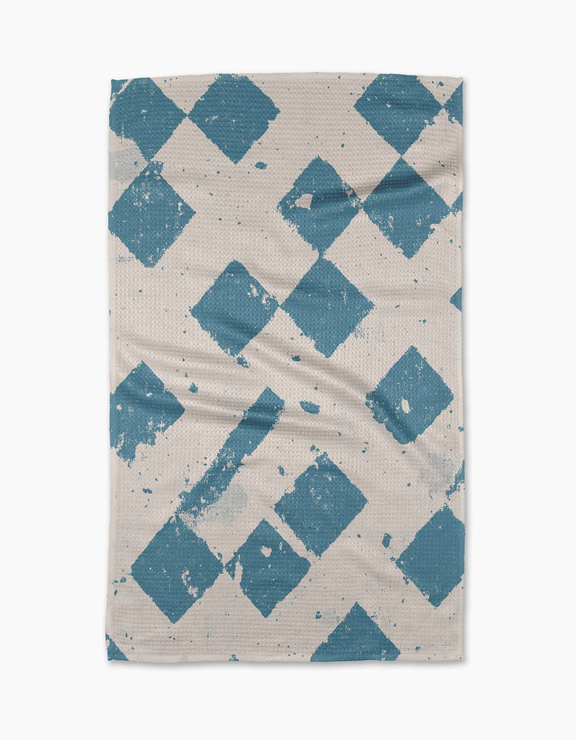 The Geometry Wisp Blue tea towel features large, distressed blue diamonds diagonally across a beige, textured surface. Some faded edges create a vintage look. Made from quick-drying, recycled materials.