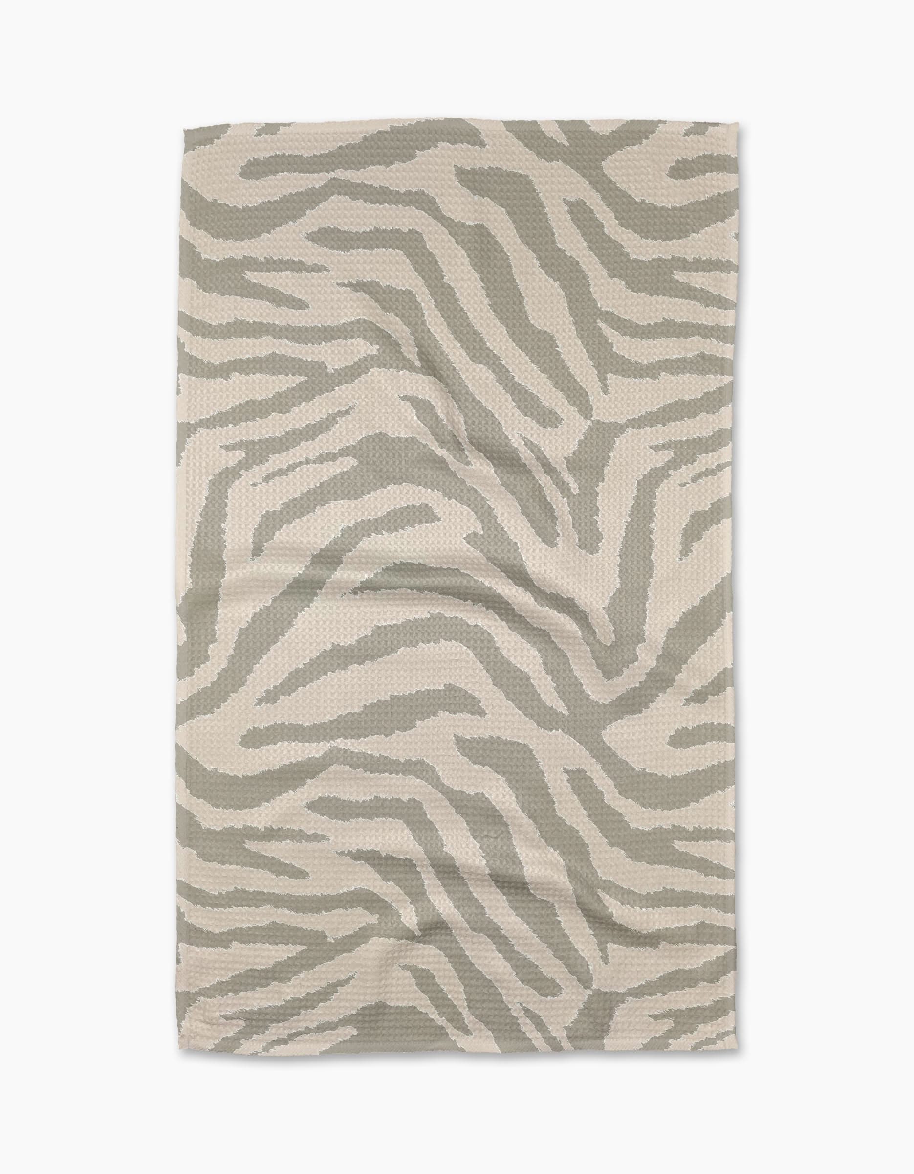The Zira Gray tea towel by Geometry is a beige, rectangular cloth made from recycled materials, featuring a subtle abstract light gray zebra stripe pattern. The towel is shown laid flat on a white background.