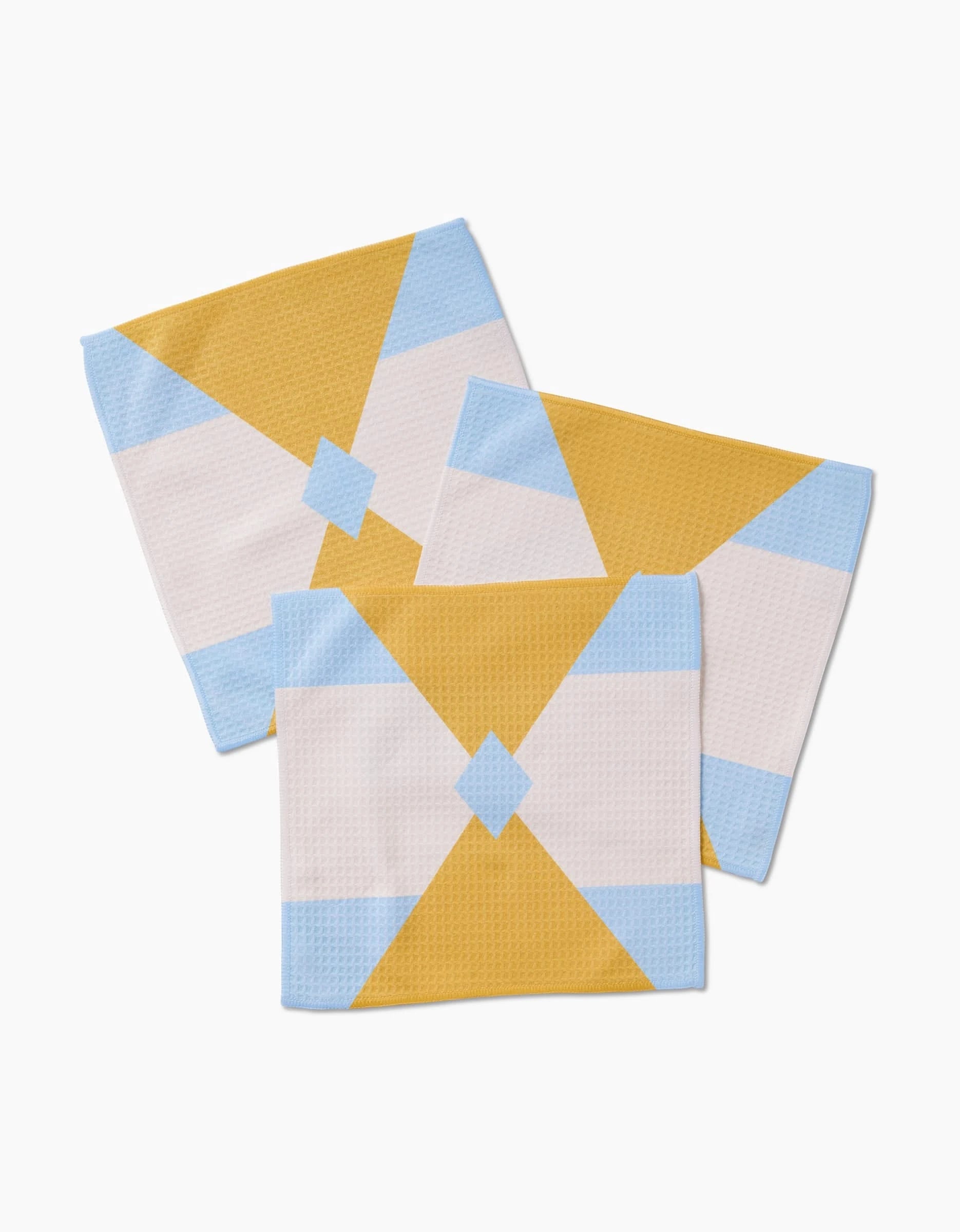 Three GEOMETRY Balanced Blue washcloths display a geometric pattern of yellow, white, and light blue triangles converging at a central blue diamond, overlapped on a plain white background.