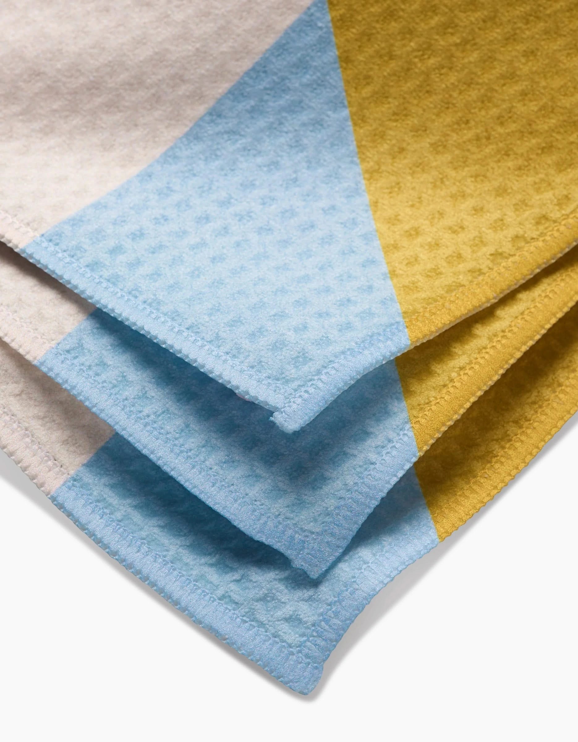 A close-up of GEOMETRYs Balanced Blue blanket showcases a geometric pattern in light blue, mustard yellow, and beige with neatly stitched edges, evoking the cozy feel of luxe washcloths.