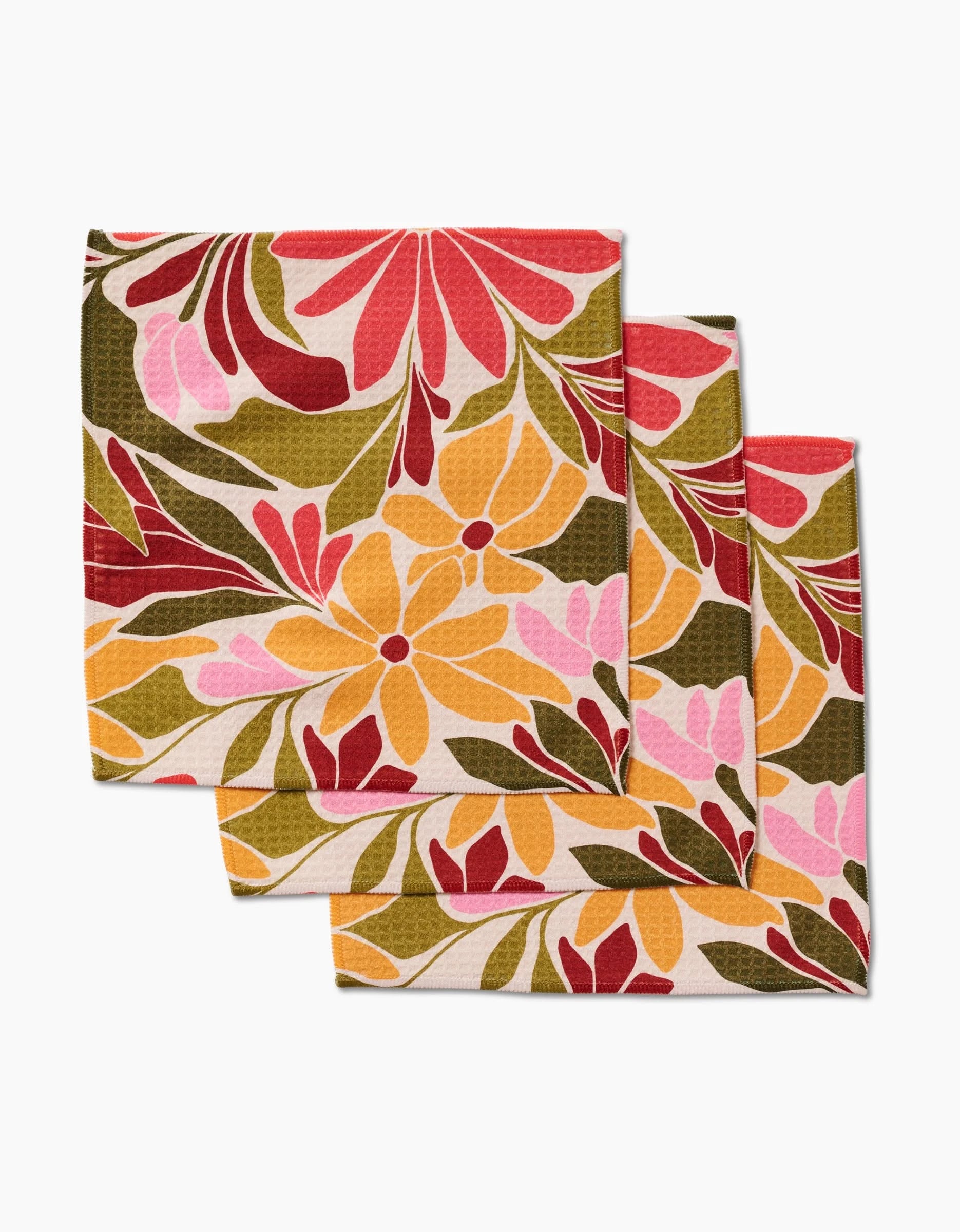 Three GEOMETRY Blooming Tan cloth napkins, featuring a floral pattern in red, pink, yellow, and green on cream recycled fabric, are arranged overlapping on a white surface.