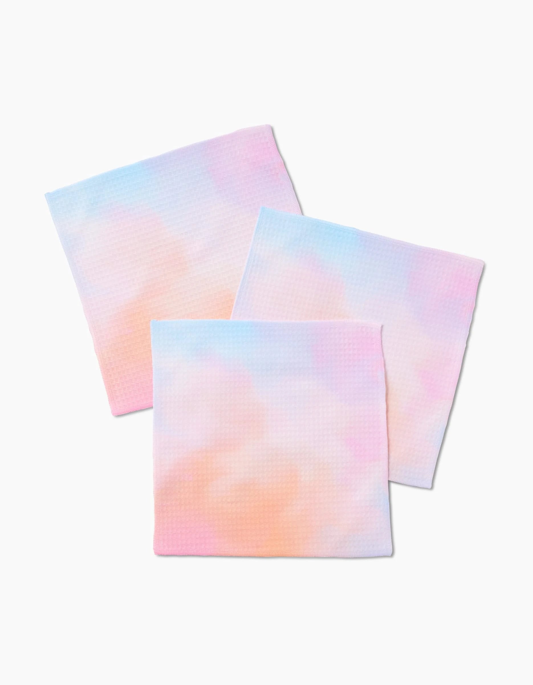 Three GEOMETRY Cotton Candy Skies Luxe Washcloths in pastel pink, blue, and peach tie-dye overlap on a white background. Made from ultra-absorbent material, they combine style and function.