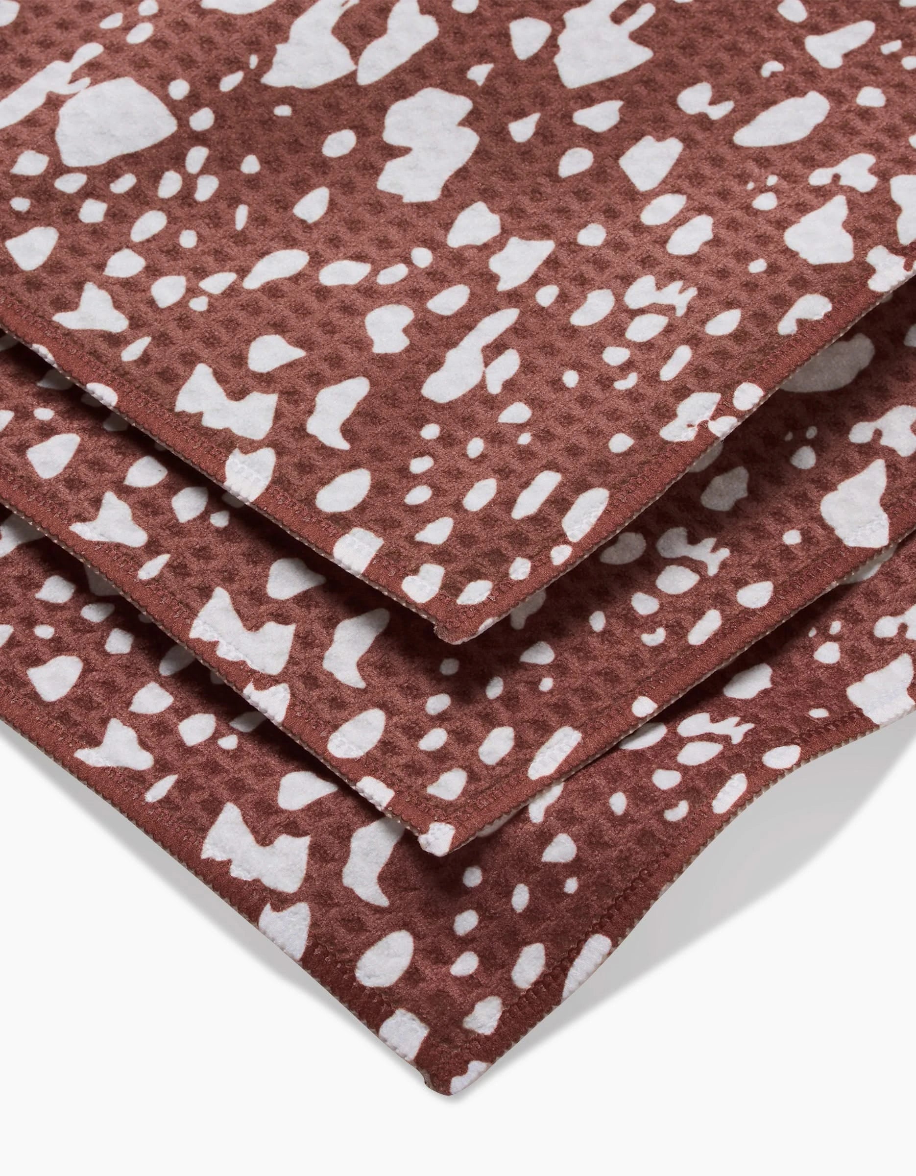 Three stacked Cowboy Horse Hide Luxe Washcloths by Hufton Studio, crafted with Geoweave technology, showcase brown fabric with irregular white spots like animal hide. Ultra-absorbent and stylish for any space, shown close up on a white background.