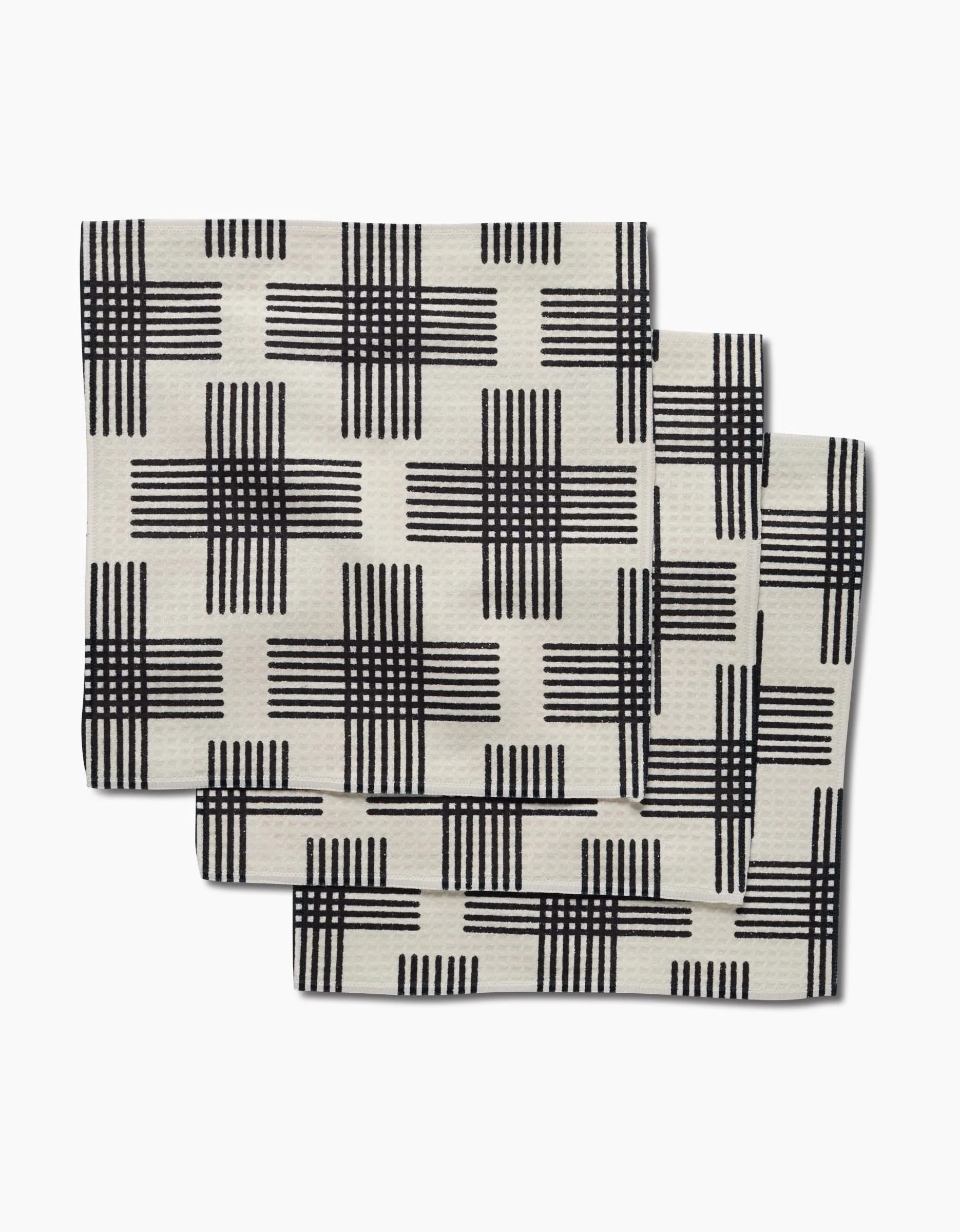 Three folded Cross Hatch napkins by Rosa Kichline, featuring a cream color with black crisscross grid and crafted with Geoweave technology for ultra-absorbency, are stacked slightly overlapping on a white background.