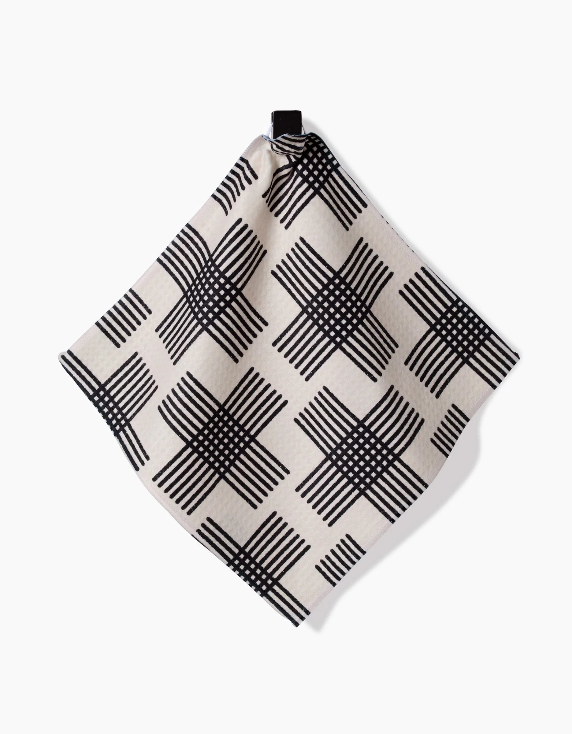 The Rosa Kichline Cross Hatch is a beige cloth napkin with a black crisscross grid, made with ultra-absorbent Geoweave fabric, hanging from a black fabric loop against a white background.