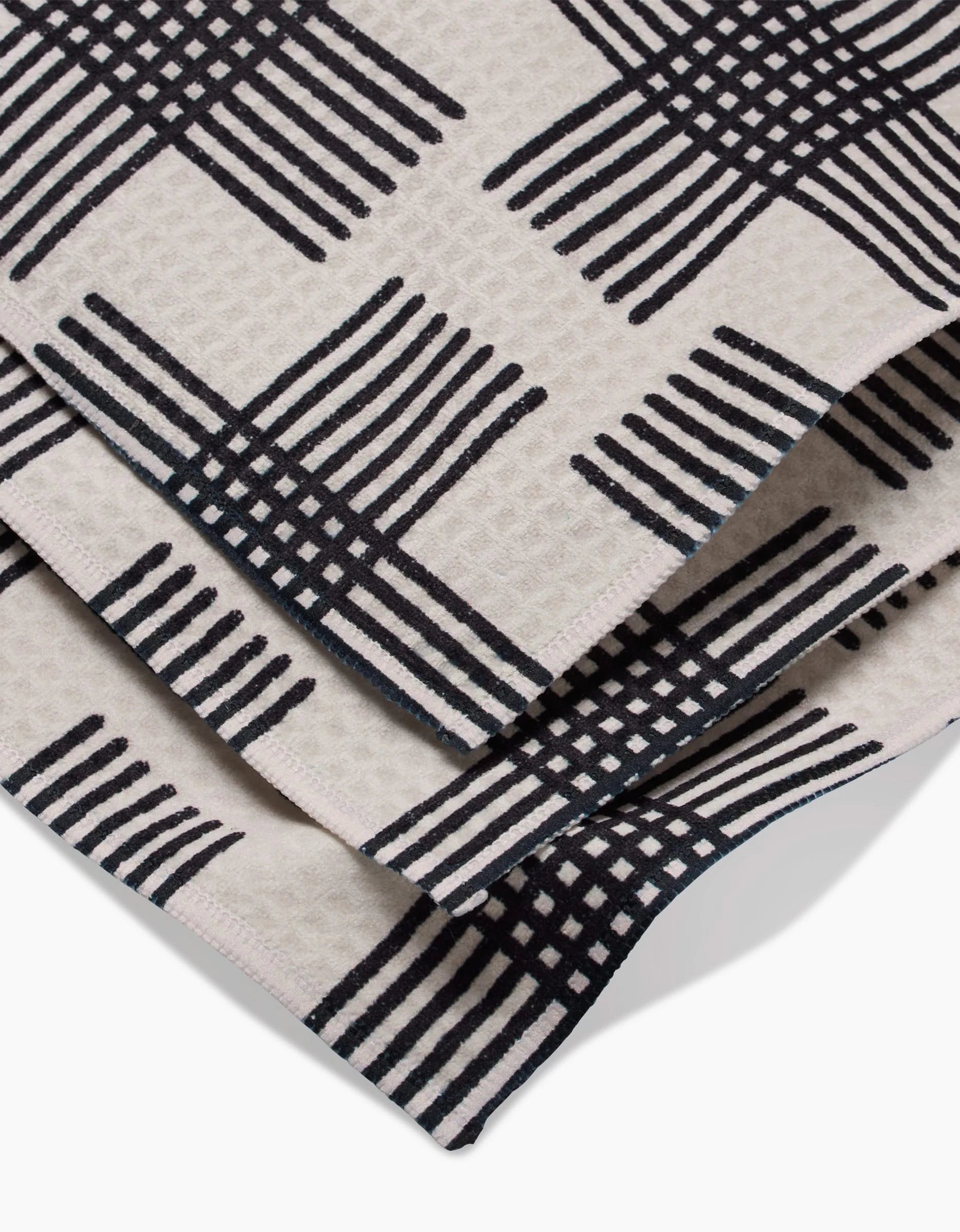 A close-up of folded beige fabric with a bold black crisscross grid, the Cross Hatch Luxe Washcloth by Rosa Kichline features a modern geometric design and ultra-absorbent Geoweave technology.
