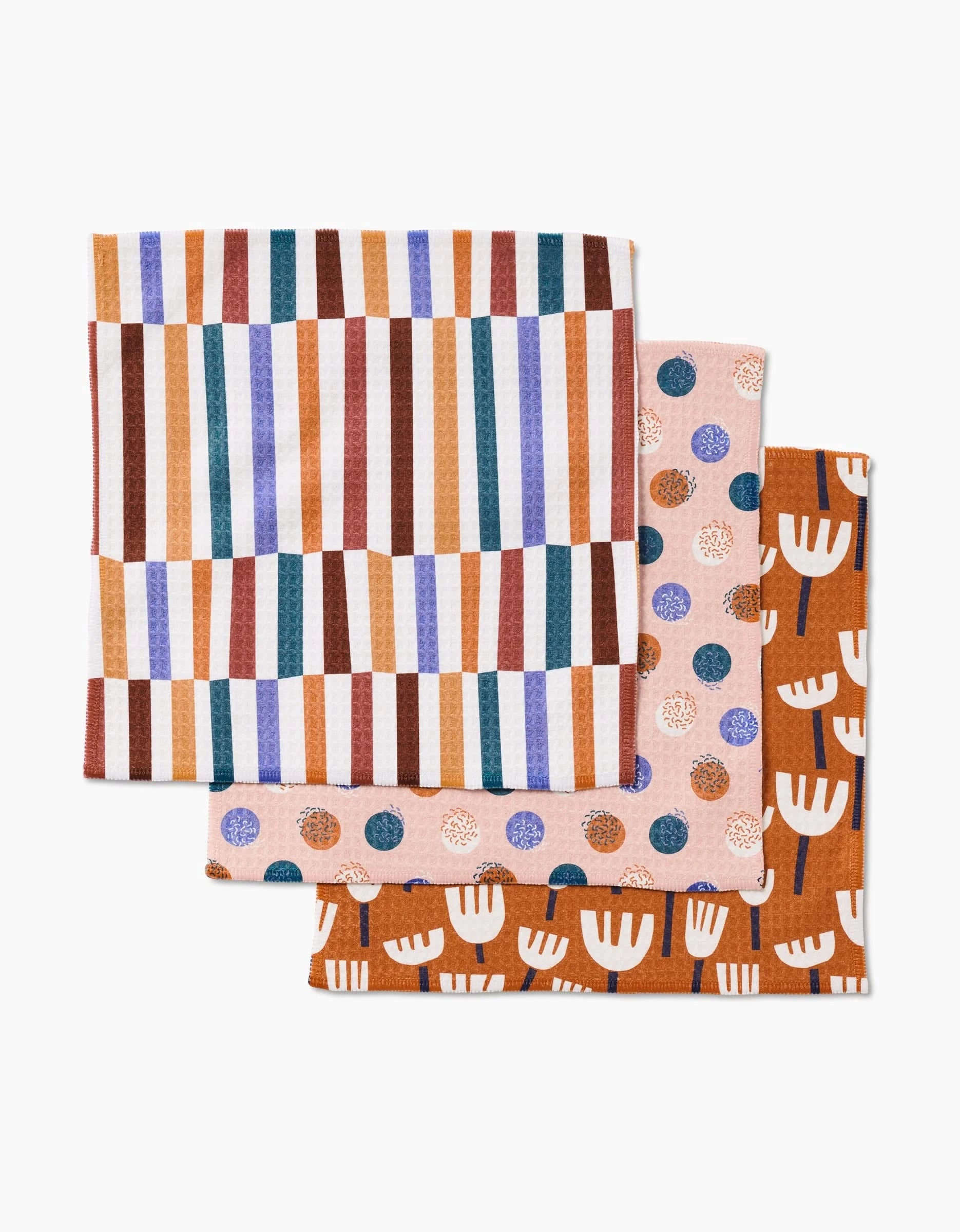 Three folded cloth napkins: one with vertical multicolored stripes, one with blue and orange polka dots on pale fabric, and Claire Iglesias’s Cutout Shapes napkin featuring white abstracts on a rust background.