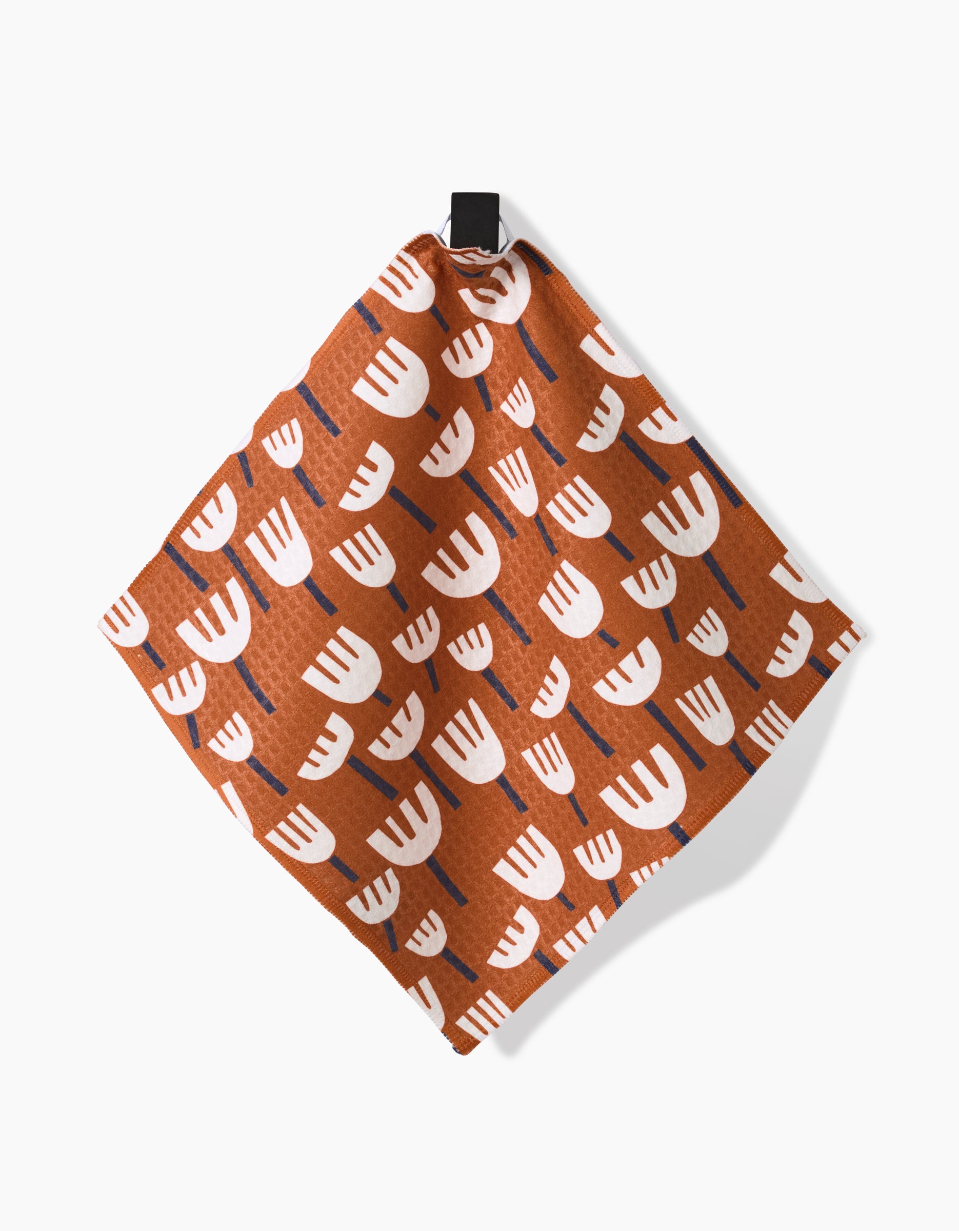 The Claire Iglesias Cutout Shapes Luxe Washcloth napkin is square and brown, with a white and blue tulip-like pattern, made from ultra-absorbent Geoweave technology. It hangs from a black loop against a white background.