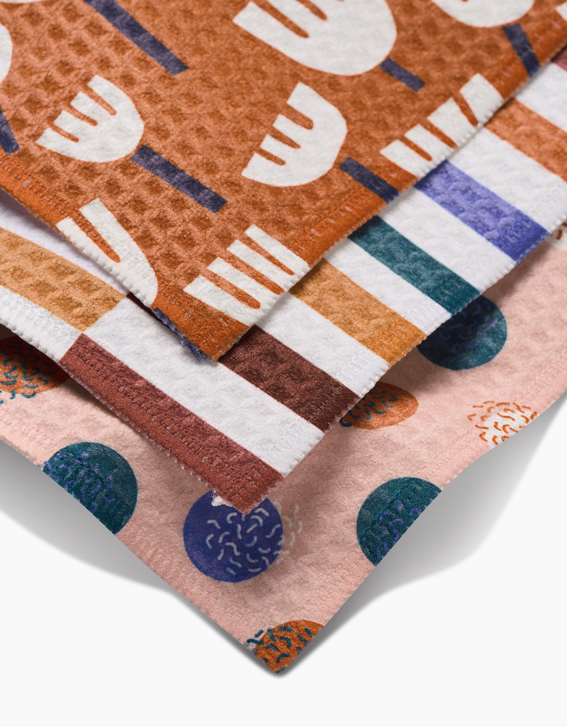 Three overlapping Claire Iglesias dish towels: Cutout Shapes (brown with white abstracts), one striped (orange, brown, blue), and one pink with blue/green circles—all made with ultra-absorbent Geoweave technology.