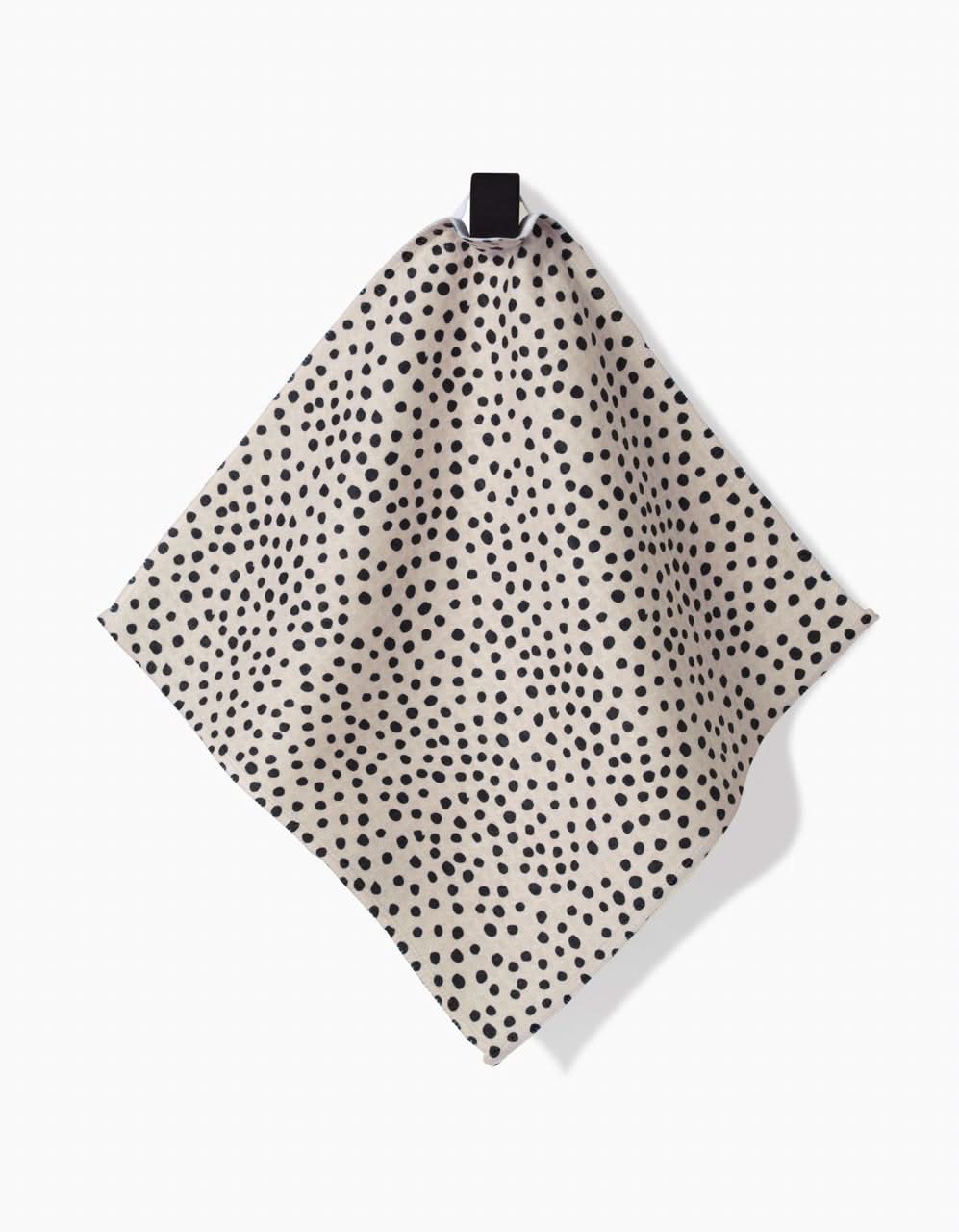 The Dotty Tan washcloth by GEOMETRY, featuring an irregular black polka dot pattern on beige and ultra-absorbent Geoweave technology, hangs from a black loop against a white background.