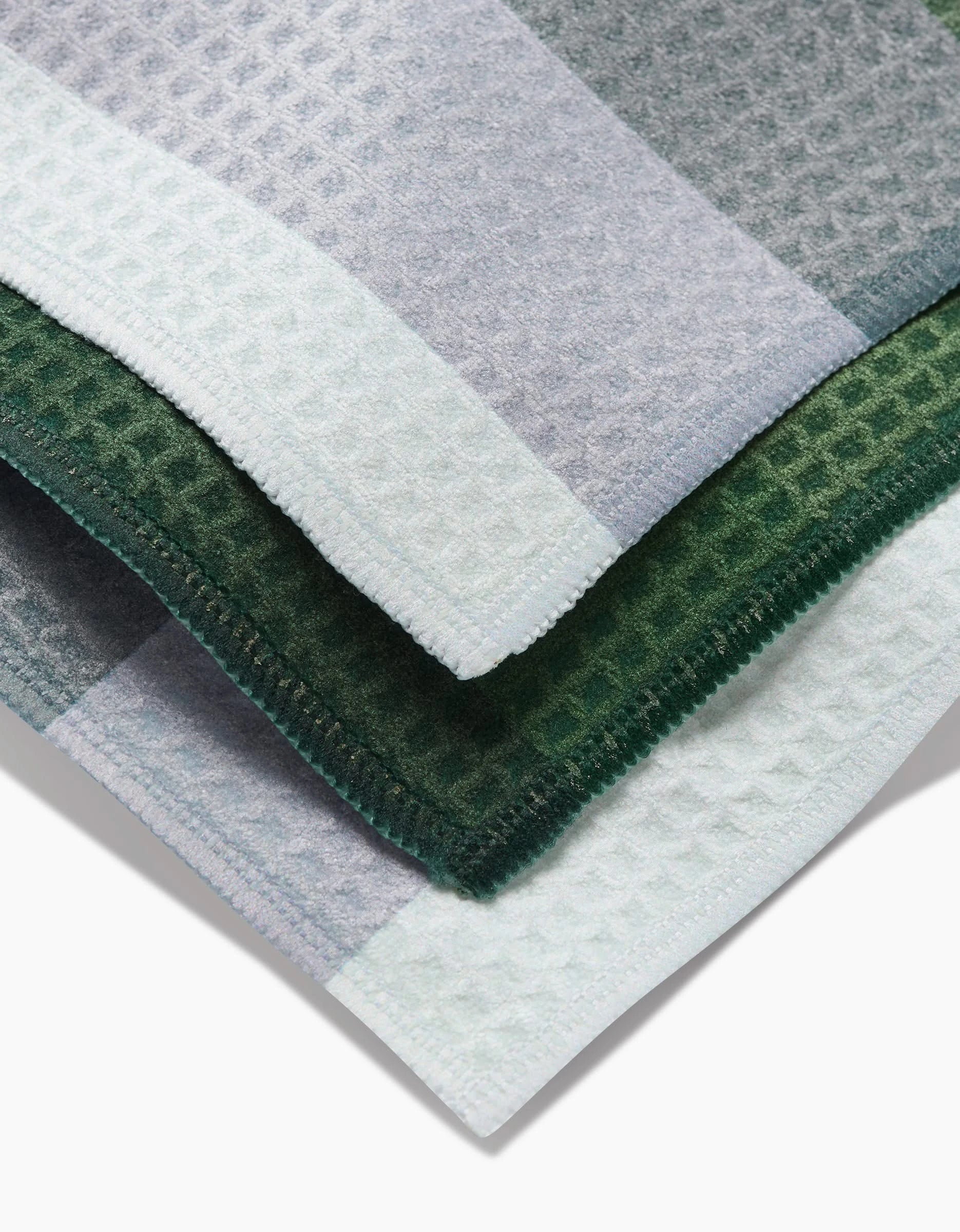 A close-up of overlapping GEOMETRY Grassy Levels washcloths in green, gray, and white highlights their ultra-absorbent Geoweave fabric and neatly stitched edges.
