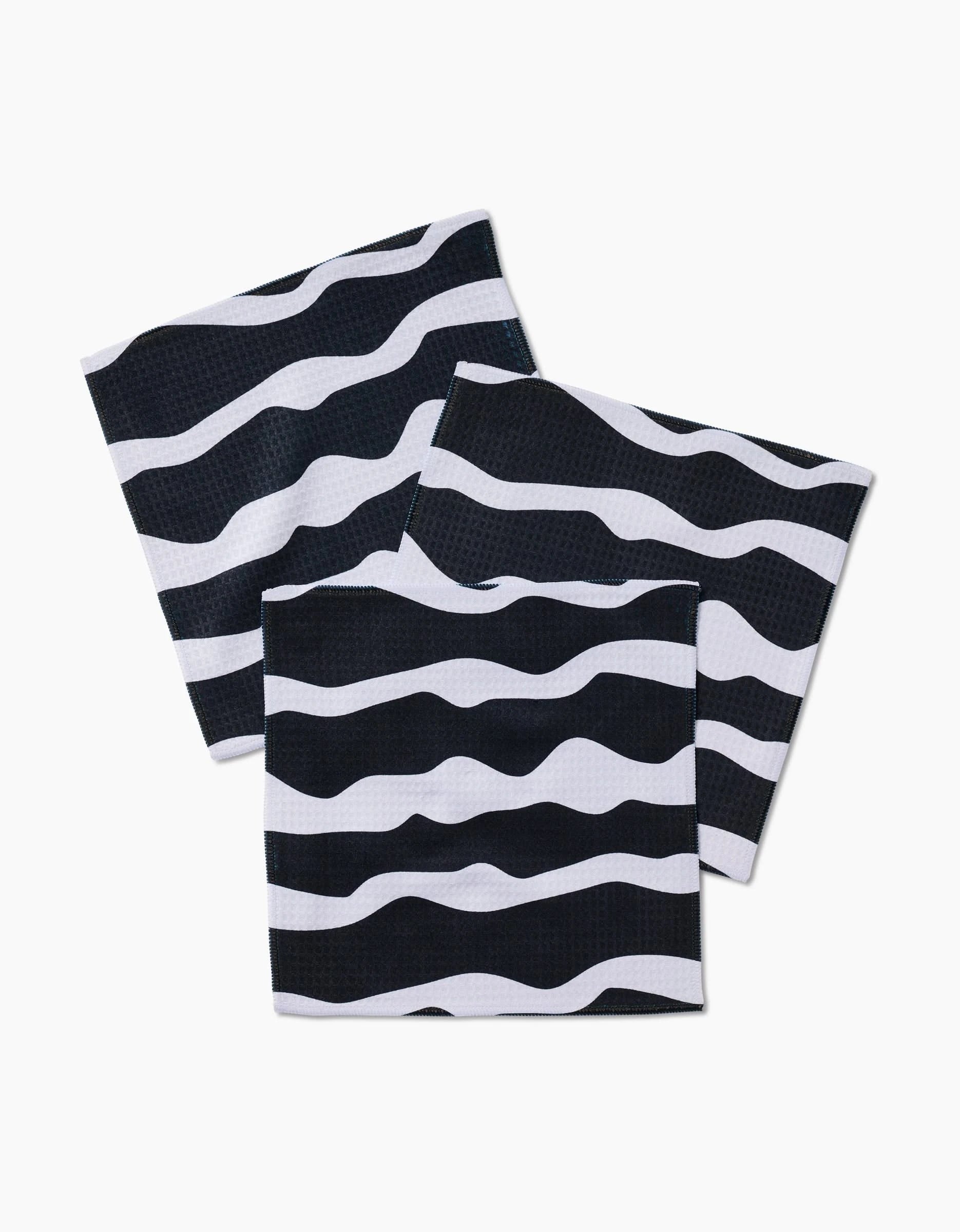Three GEOMETRY Ink Twist black square napkins, featuring wavy white stripes and made from recycled materials, are arranged in an overlapping pattern on a white background.
