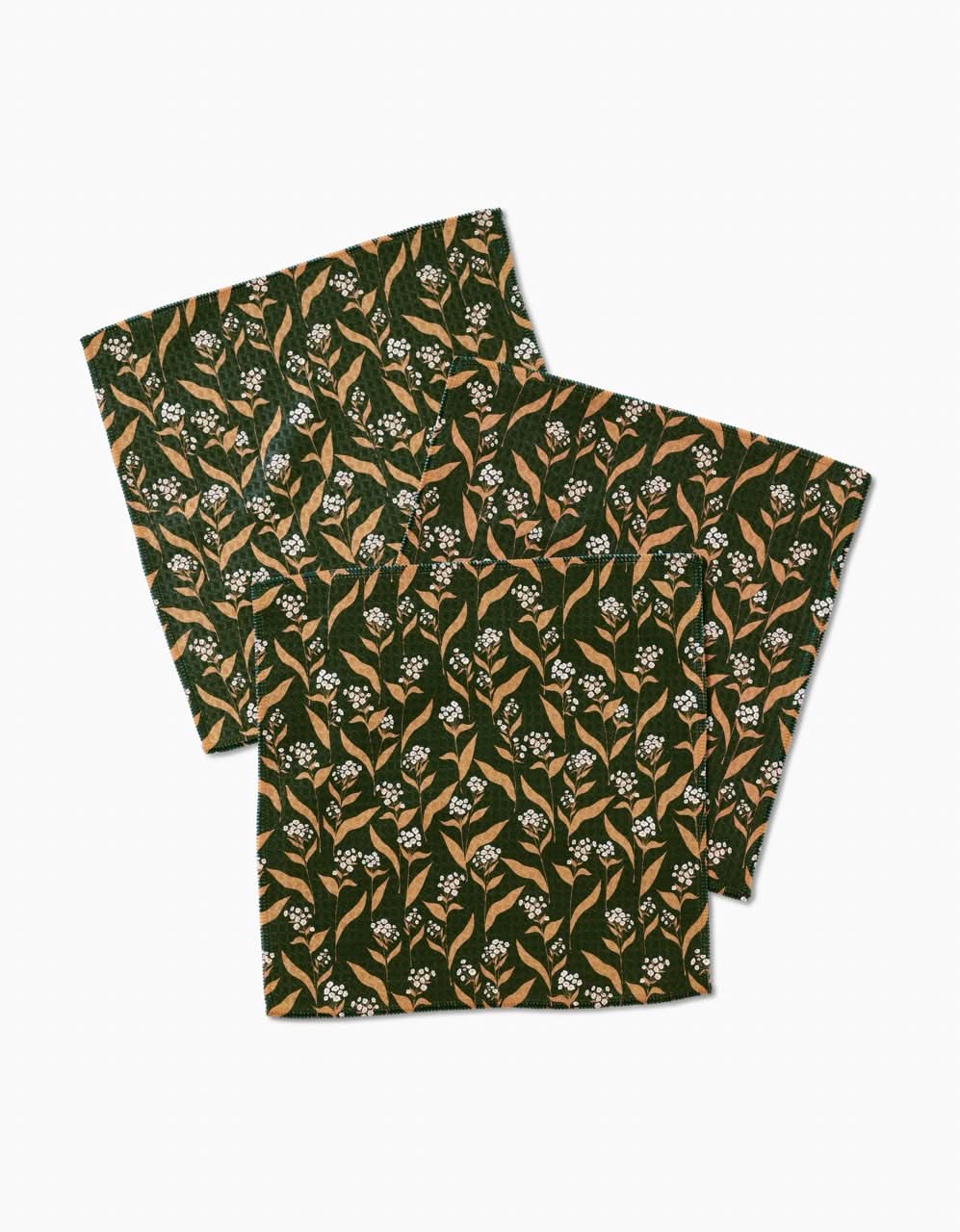 The Loretta napkin set by Charlie Rowan Designs includes two luxe square cloth napkins with a dark green background, tan leaves, and white floral prints—perfect for an elegant table setting.