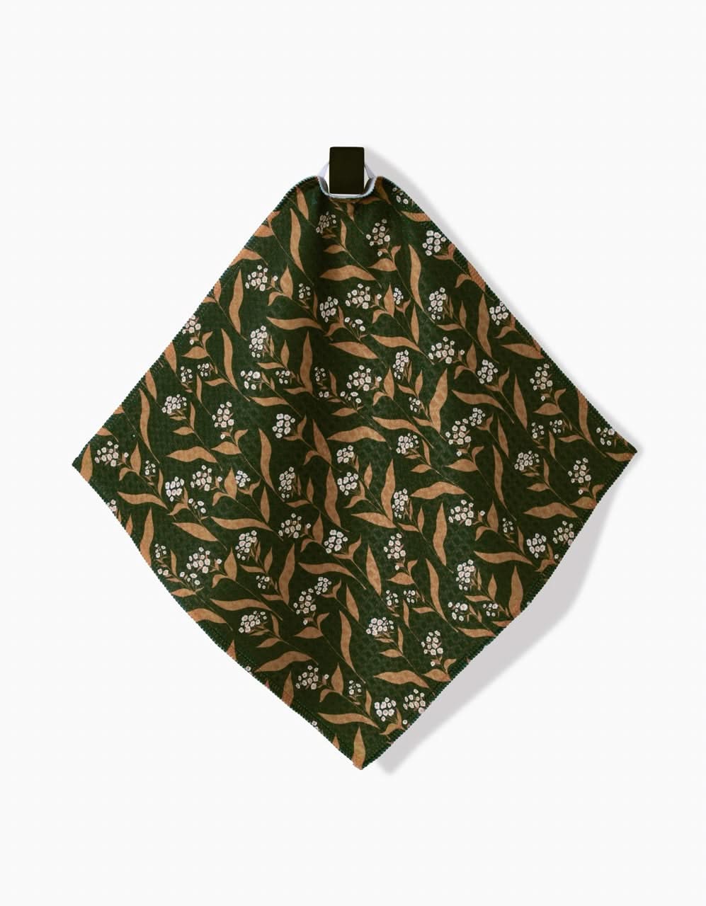 The Loretta napkin by Charlie Rowan Designs is a square green cloth made from recycled materials, featuring brown branches, tan leaves, and white flowers. Its shown hanging from a metal clip against a white background.