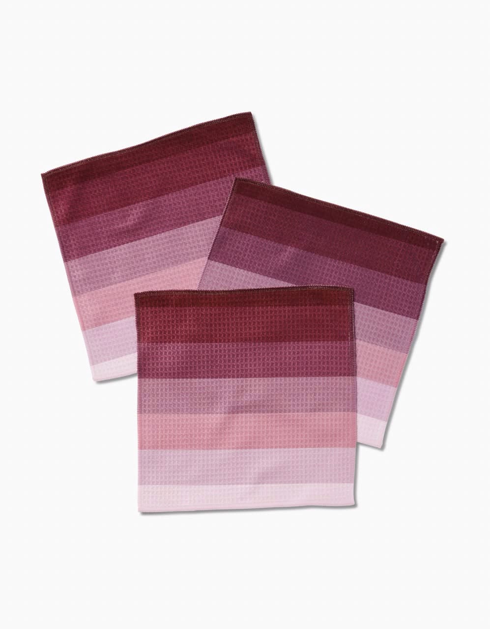 Three folded GEOMETRY Mauve Levels Luxe Washcloth napkins, with horizontal stripes in pink, red, and burgundy, overlap on a white background. Made with ultra-absorbent Geoweave technology for superior performance.