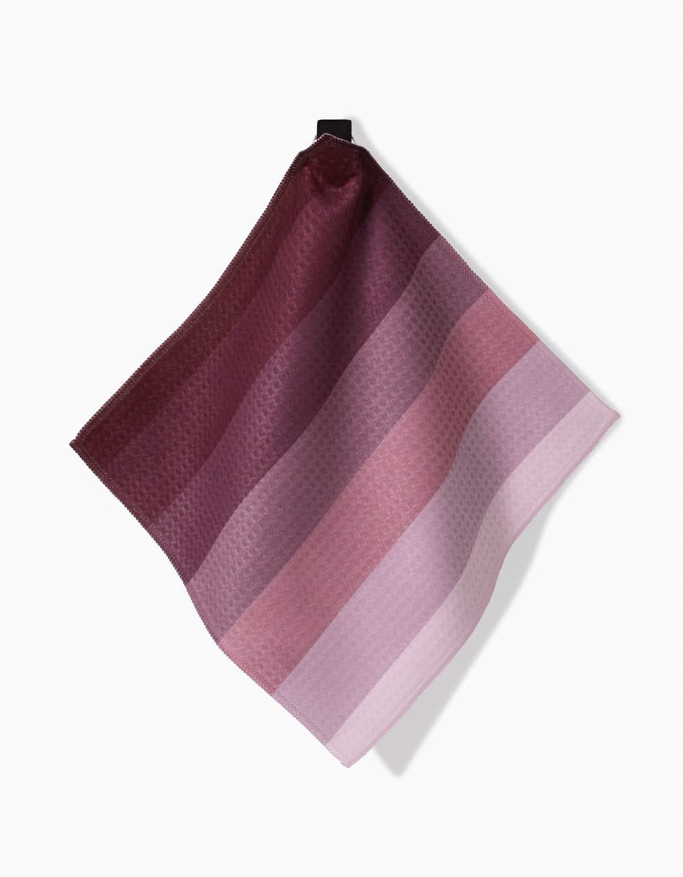 The GEOMETRY Mauve Levels kitchen towel, made with Geoweave technology, displays burgundy, mauve, and pink horizontal stripes and hangs from a black loop against a white background.