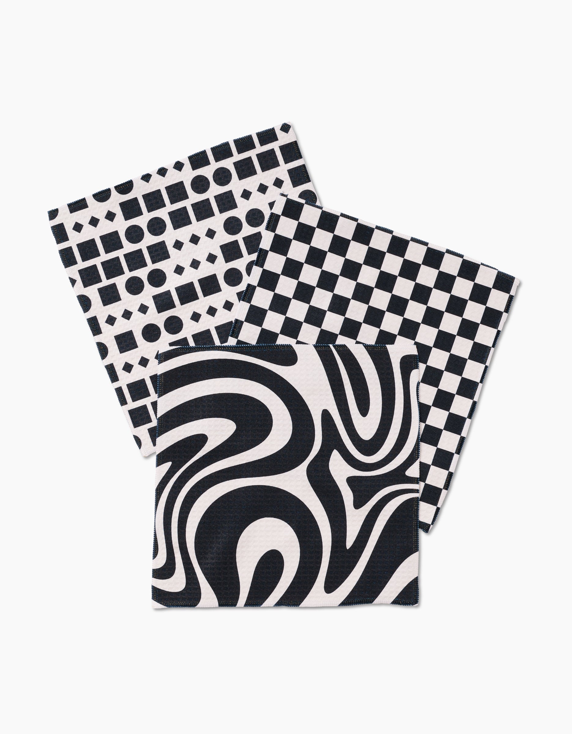 Three GEOMETRY Luxe Mixed Up washcloths—featuring grid, checkered, and swirling black-and-white patterns—are stacked in layers on a white background.