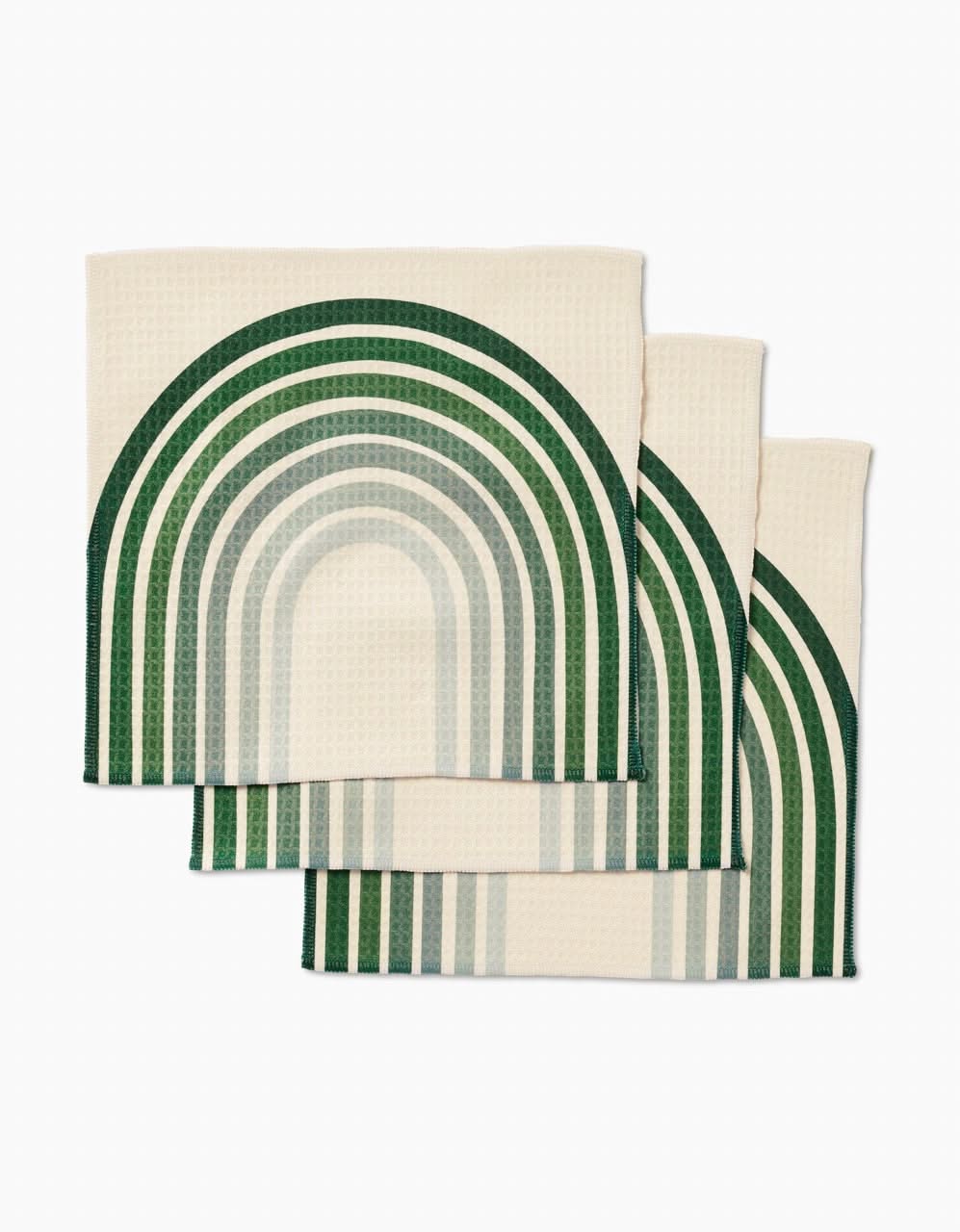 Three cream-colored GEOMETRY Olive Arches Luxe Washcloths, featuring a green and teal geometric rainbow pattern, are neatly stacked with overlapping curved designs against a white background.