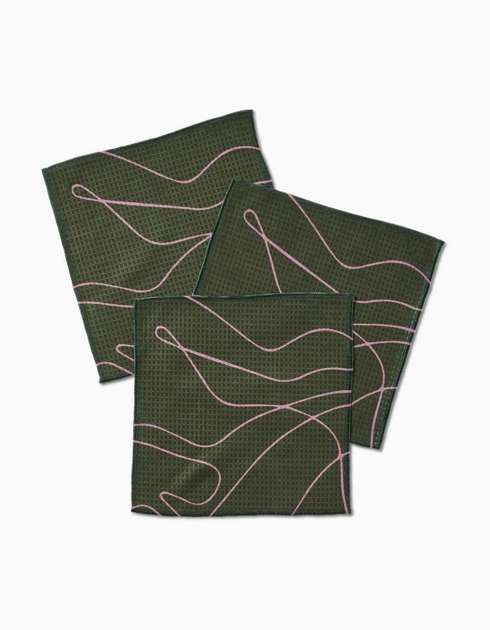 Three GEOMETRY Olive Martini napkins, featuring a textured dark green pattern and wavy pink lines, are overlapped on a white background. Made with Geoweave technology, these ultra-absorbent napkins offer everyday luxury.