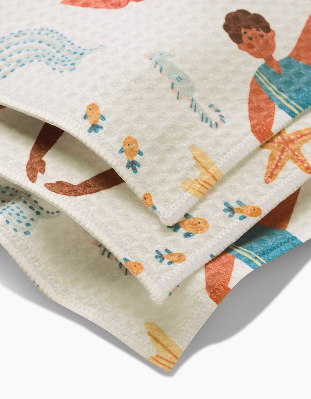 A close-up of the folded, textured Spirit of the Ocean washcloth by Janna Sue Design, featuring a playful beach-inspired print with illustrated people, starfish, fish, and waves.