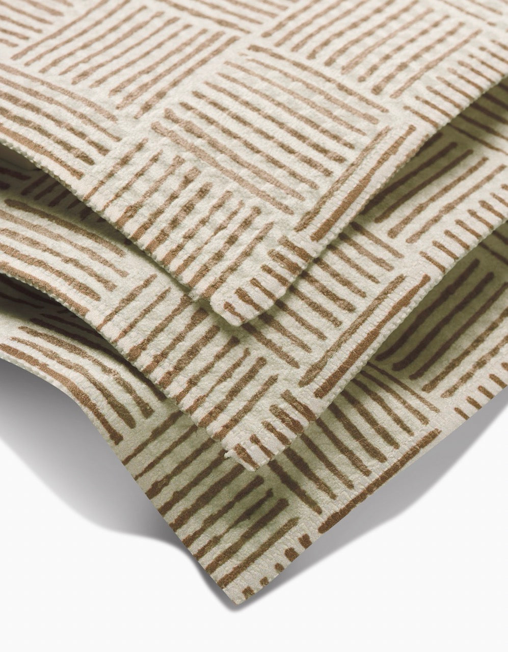Three overlapping Studio Jeannot Thatcher Luxe Washcloths made from recycled materials, with a repeating brown hand-drawn diagonal line square pattern. These ultra-absorbent sheets are neatly stacked on a white surface.