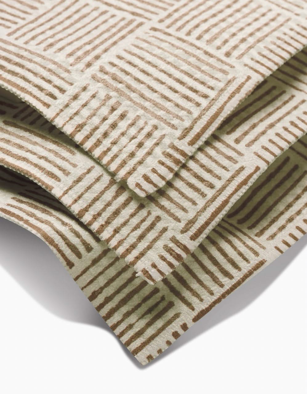 Three overlapping Studio Jeannot Thatcher Luxe Washcloths made from recycled materials, with a repeating brown hand-drawn diagonal line square pattern. These ultra-absorbent sheets are neatly stacked on a white surface.