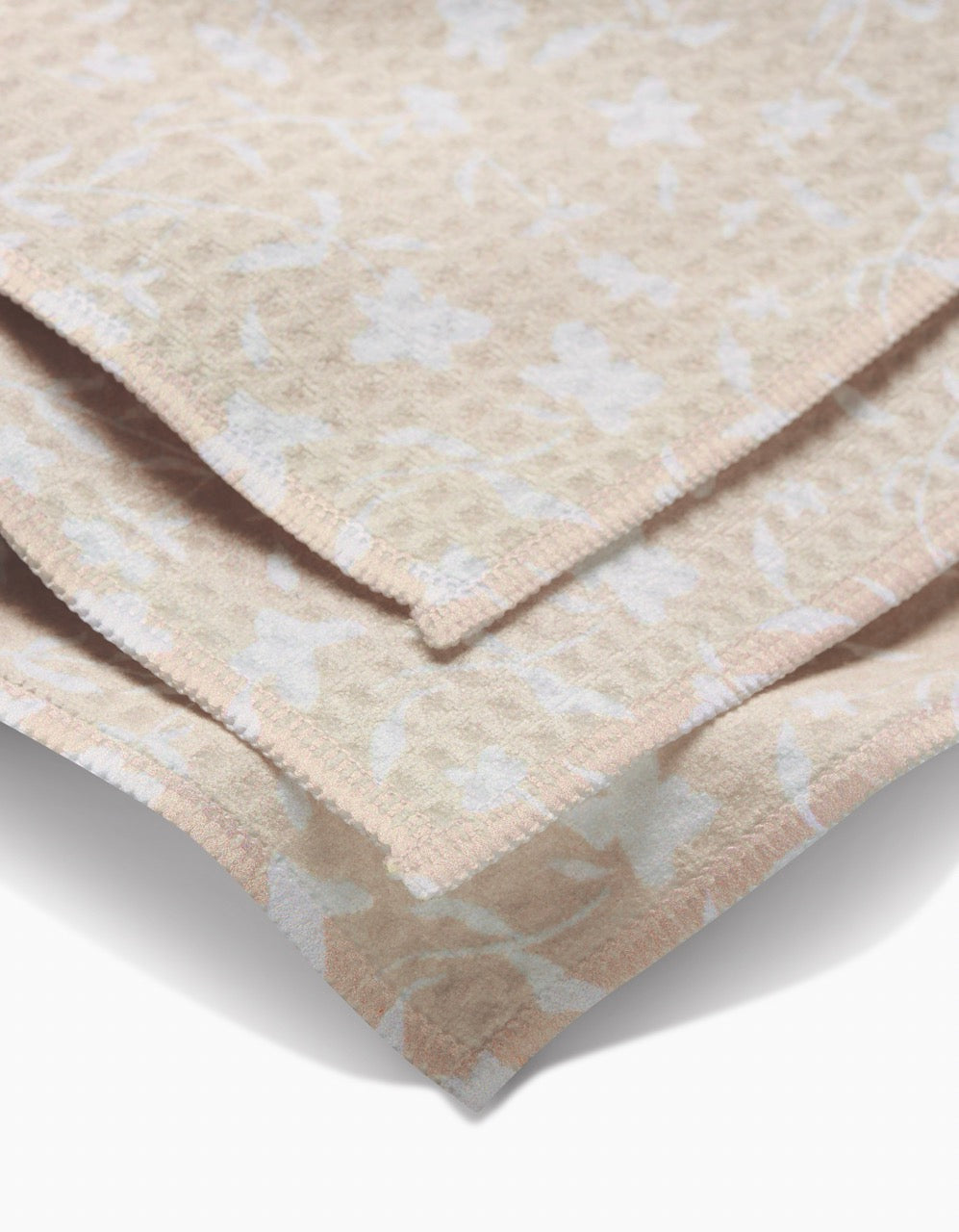Close-up of a folded Aubrey Fairchild Tiny Floral washcloth in beige and cream, featuring a subtle floral pattern. Geoweave technology provides ultra-absorbent softness and neatly finished edges for a luxurious feel.