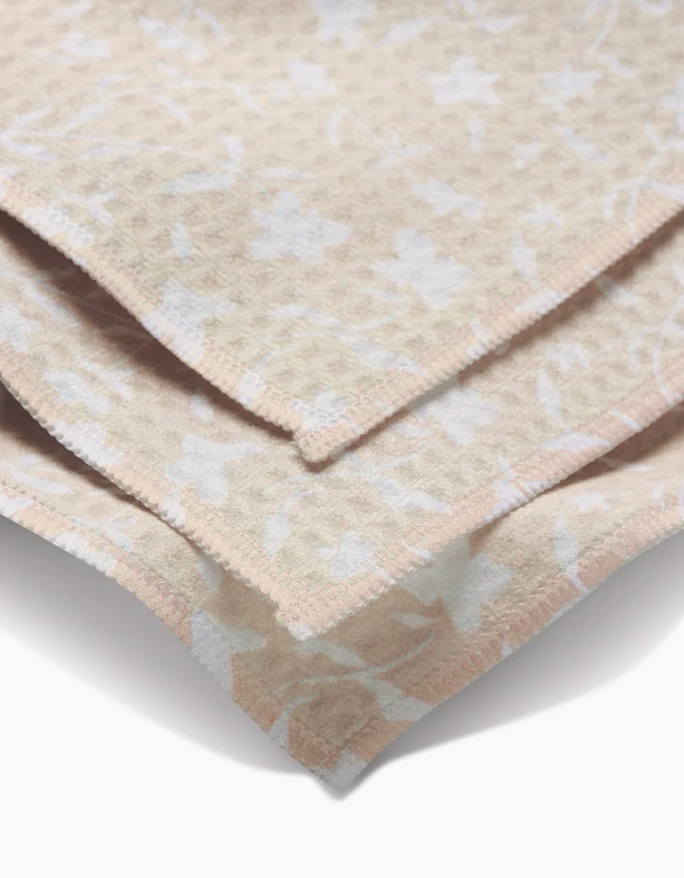 Close-up of a folded Aubrey Fairchild Tiny Floral washcloth in beige and cream, featuring a subtle floral pattern. Geoweave technology provides ultra-absorbent softness and neatly finished edges for a luxurious feel.