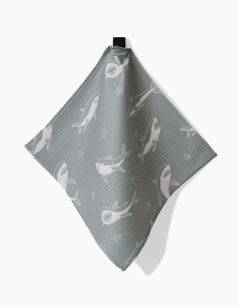 The Toothy Tales Luxe Washcloth by Janna Sue Design is a textured, square, light gray-blue cloth made from recycled materials with white shark illustrations, hanging by a black loop against a plain white background.