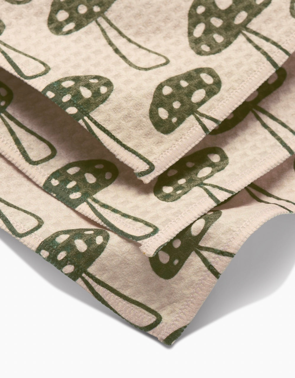 The Kayla Ann Woodland Mushroom Luxe Washcloth features beige textured fabric with a repeating green mushroom and white spot pattern. Its layered, ultra-absorbent, made from post-consumer recycled materials, and finished with neat stitching.