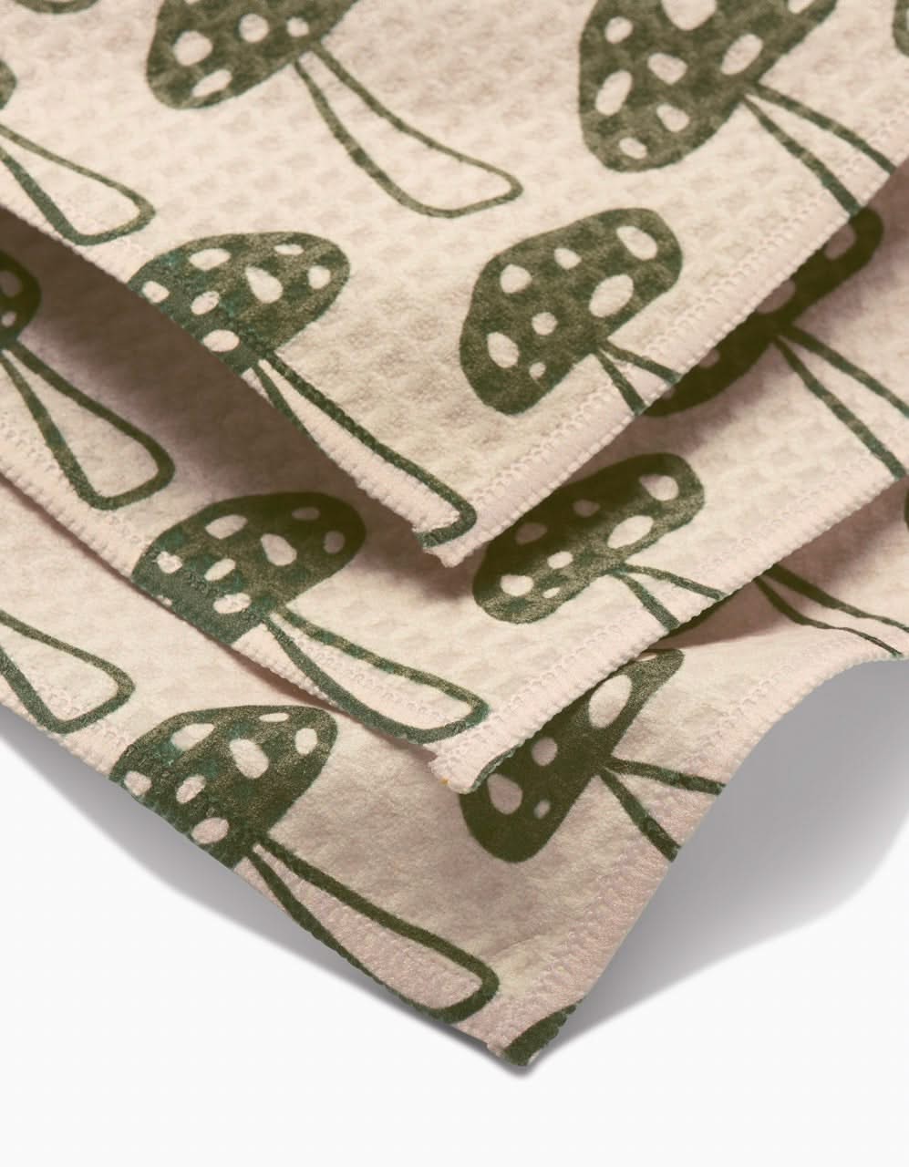The Kayla Ann Woodland Mushroom Luxe Washcloth features beige textured fabric with a repeating green mushroom and white spot pattern. Its layered, ultra-absorbent, made from post-consumer recycled materials, and finished with neat stitching.