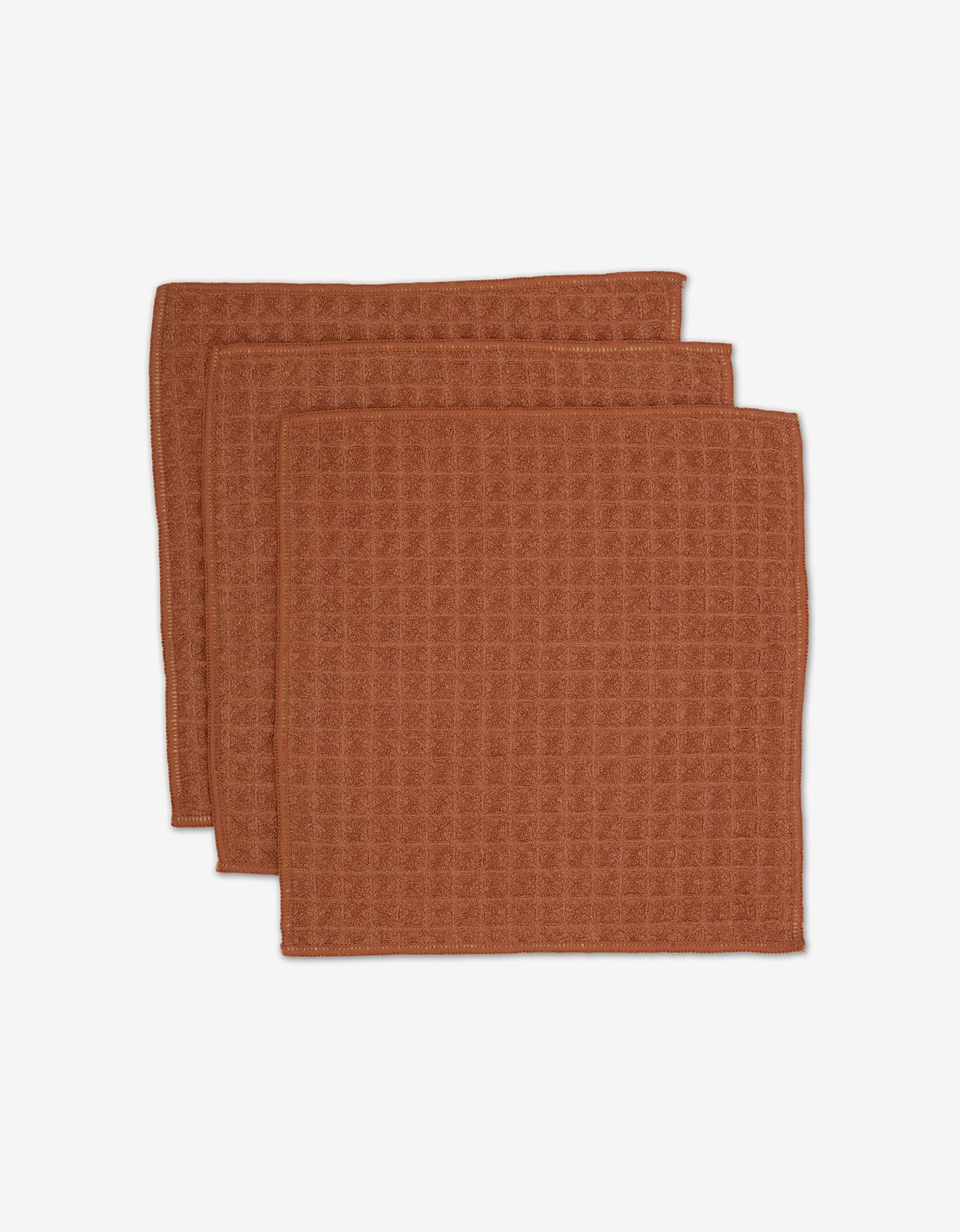 Three folded brown waffle-textured absorbent washcloths, stacked and slightly overlapping on a white background. GEOMETRY’s Clay washcloths from the Waffle Bath Collection.