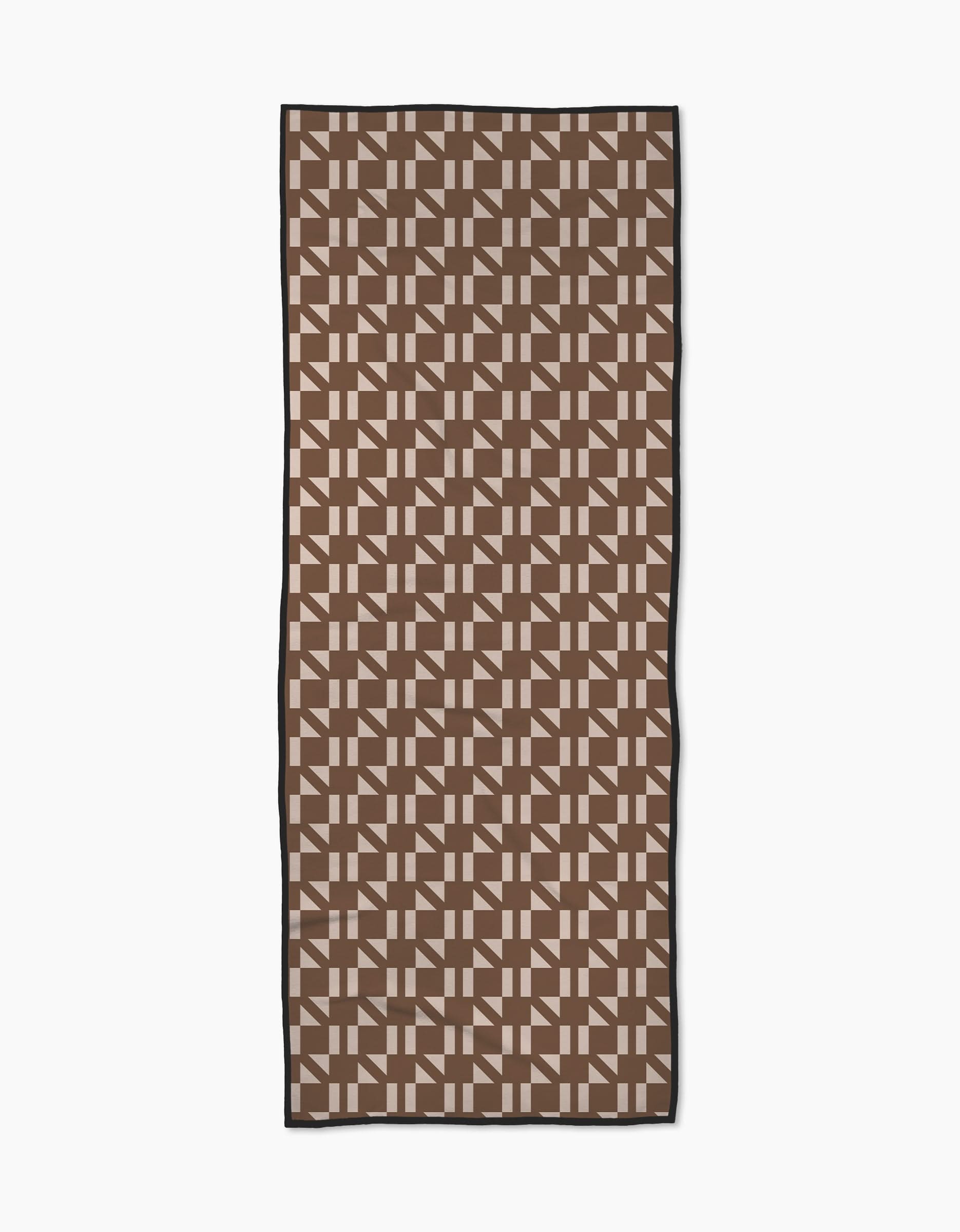 The Geometry Affogato yoga towel features a repeating geometric pattern of interlocking brown and beige shapes, framed by a thin black border. Made from recycled materials, it’s shown on a white background.