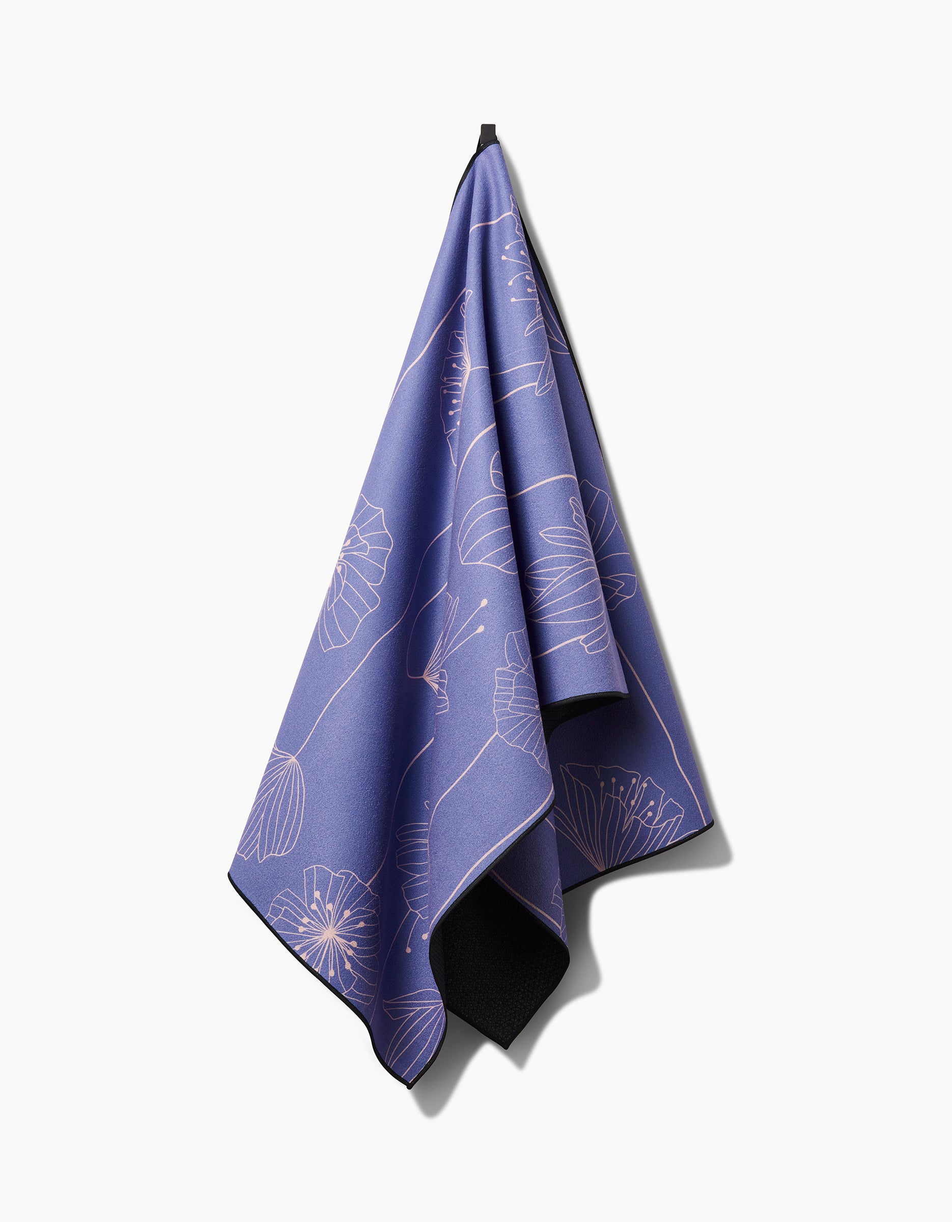 The GEOMETRY Alexa yoga towel, featuring a light abstract floral pattern in purple, hangs from a loop against a white background. Made with recycled materials, it casts a gentle shadow to the right.
