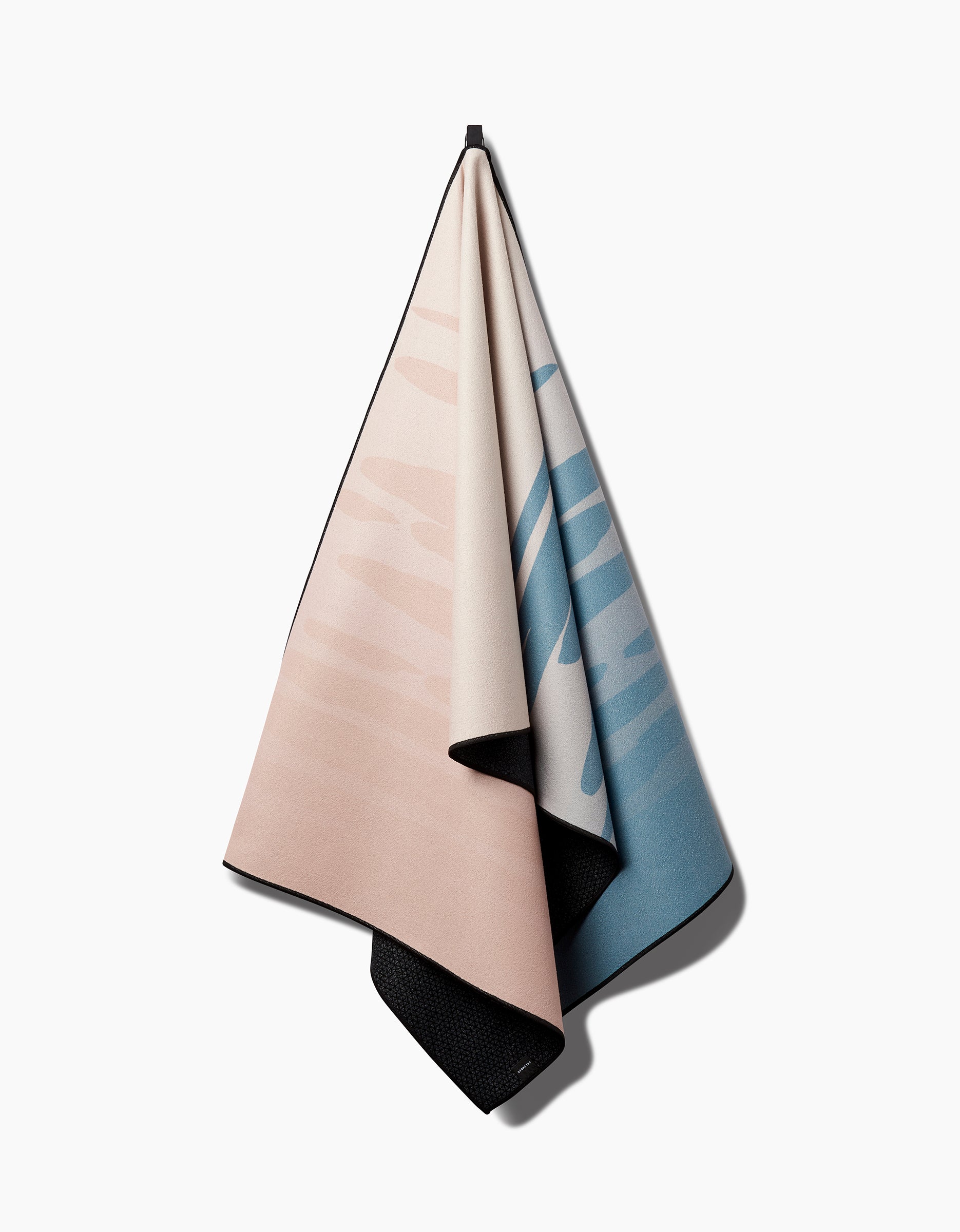 The GEOMETRY Balance yoga towel, featuring a slip-resistant surface and a soft gradient from light beige to blue, hangs neatly from a hook against a plain white background.