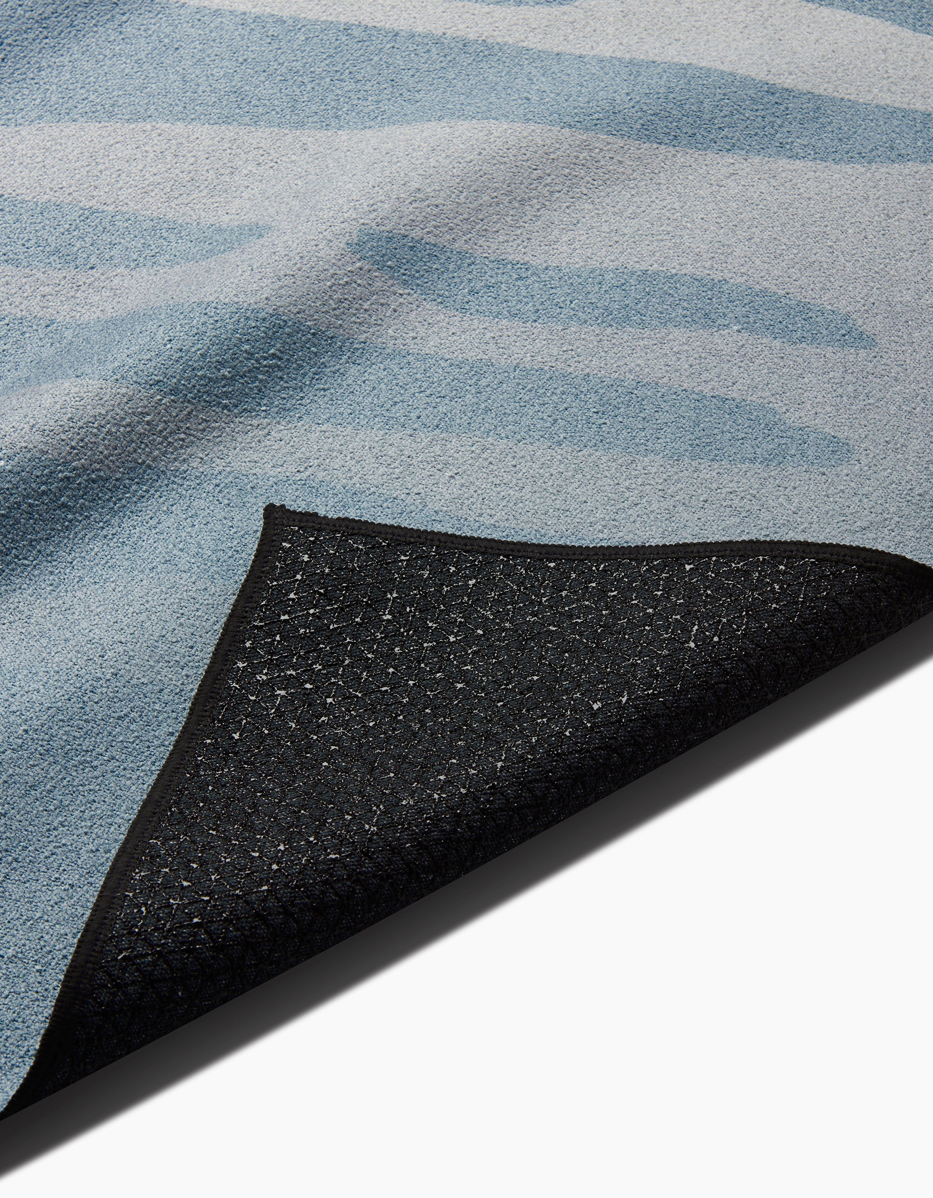 A close-up of the GEOMETRY Balance rug shows its blue pattern and a flipped corner, revealing a textured black non-slip backing inspired by slip-resistant yoga towel materials.