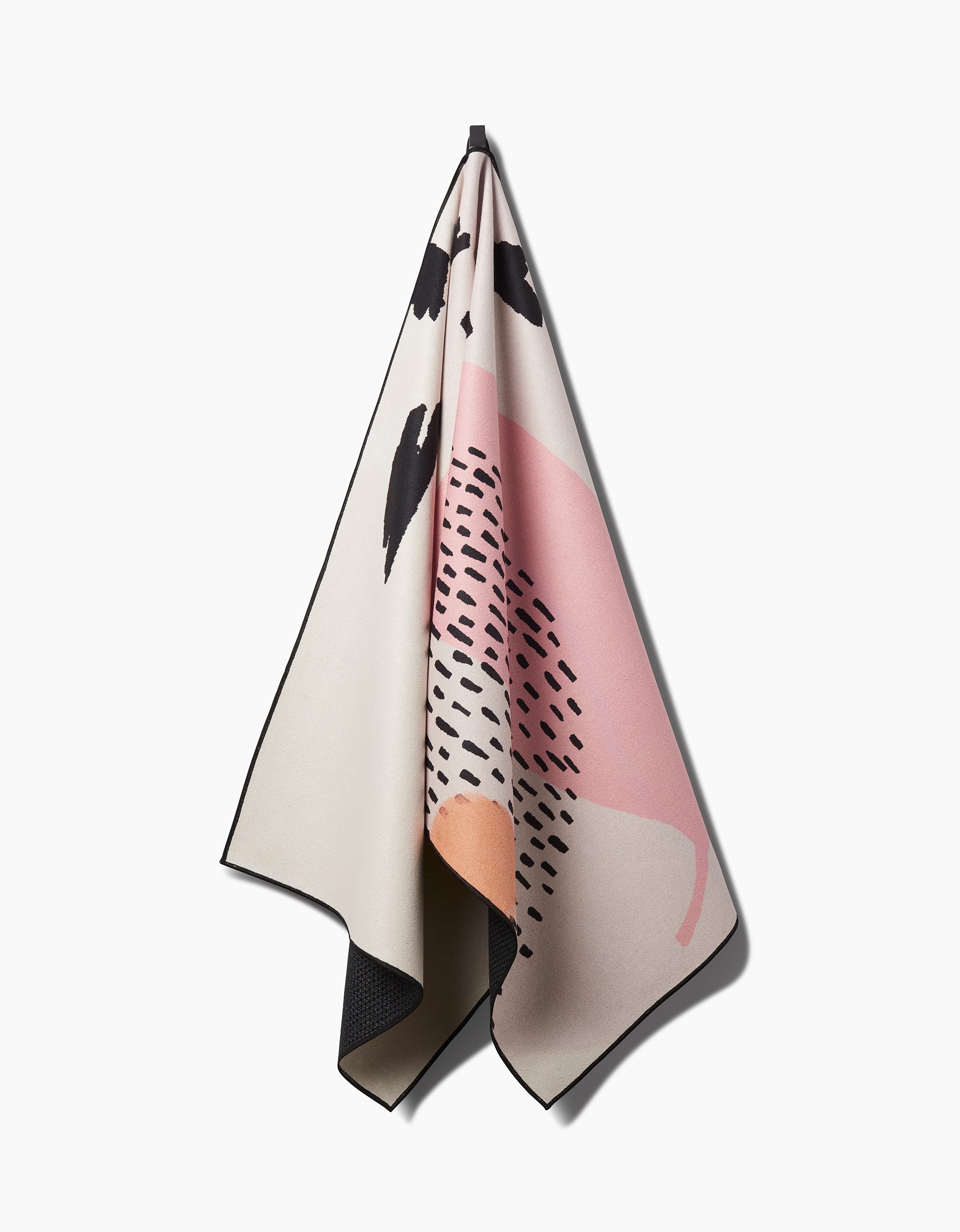 The GEOMETRY Bare recycled yoga towel in beige, featuring abstract black, pink, and orange shapes and patterns, hangs from a hook against a white background.