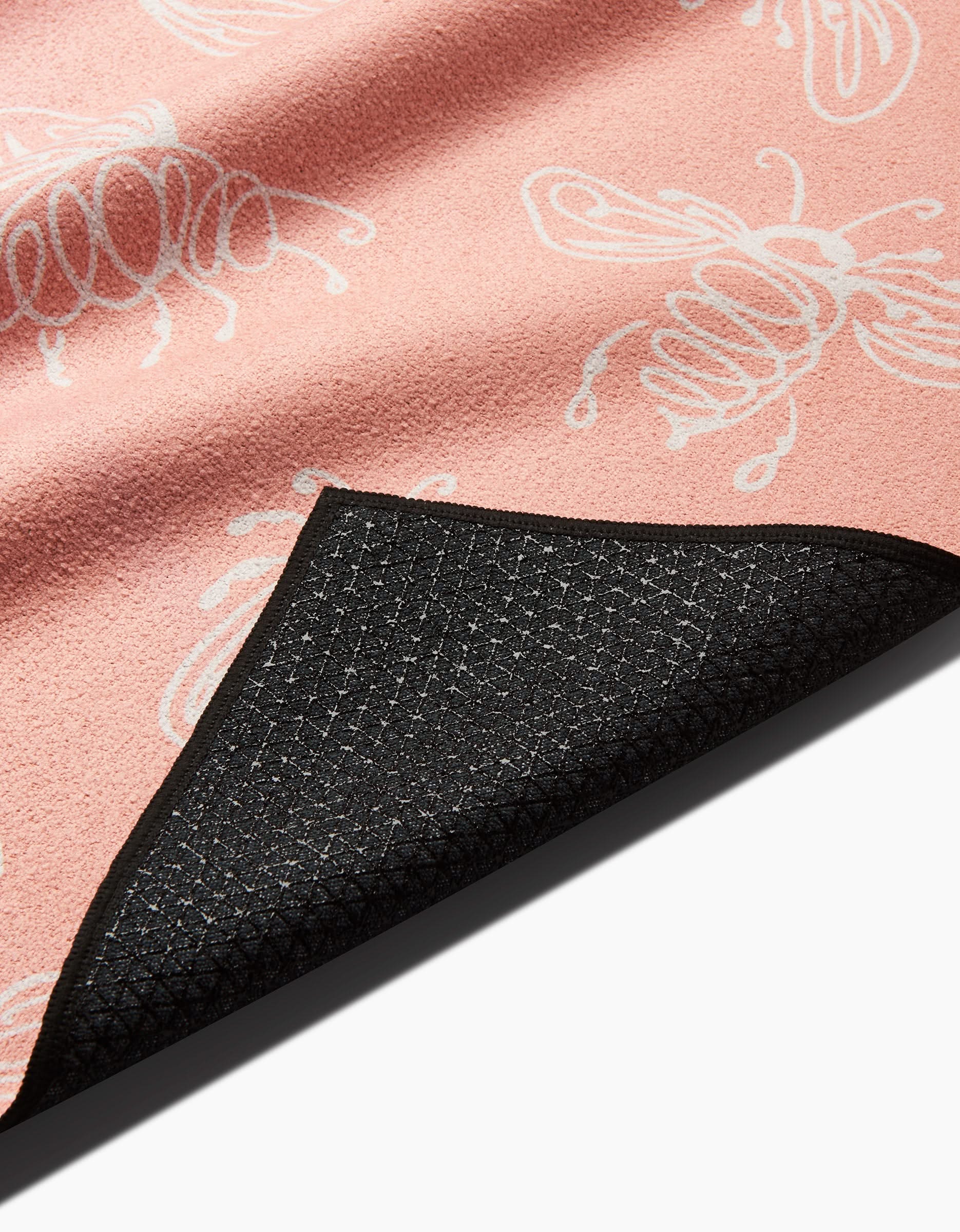 A close-up of the Sara Mari Beelieve pink yoga towel with white bee illustrations, showcasing its textured black non-slip backing partly folded to reveal the underside of this recycled towel.