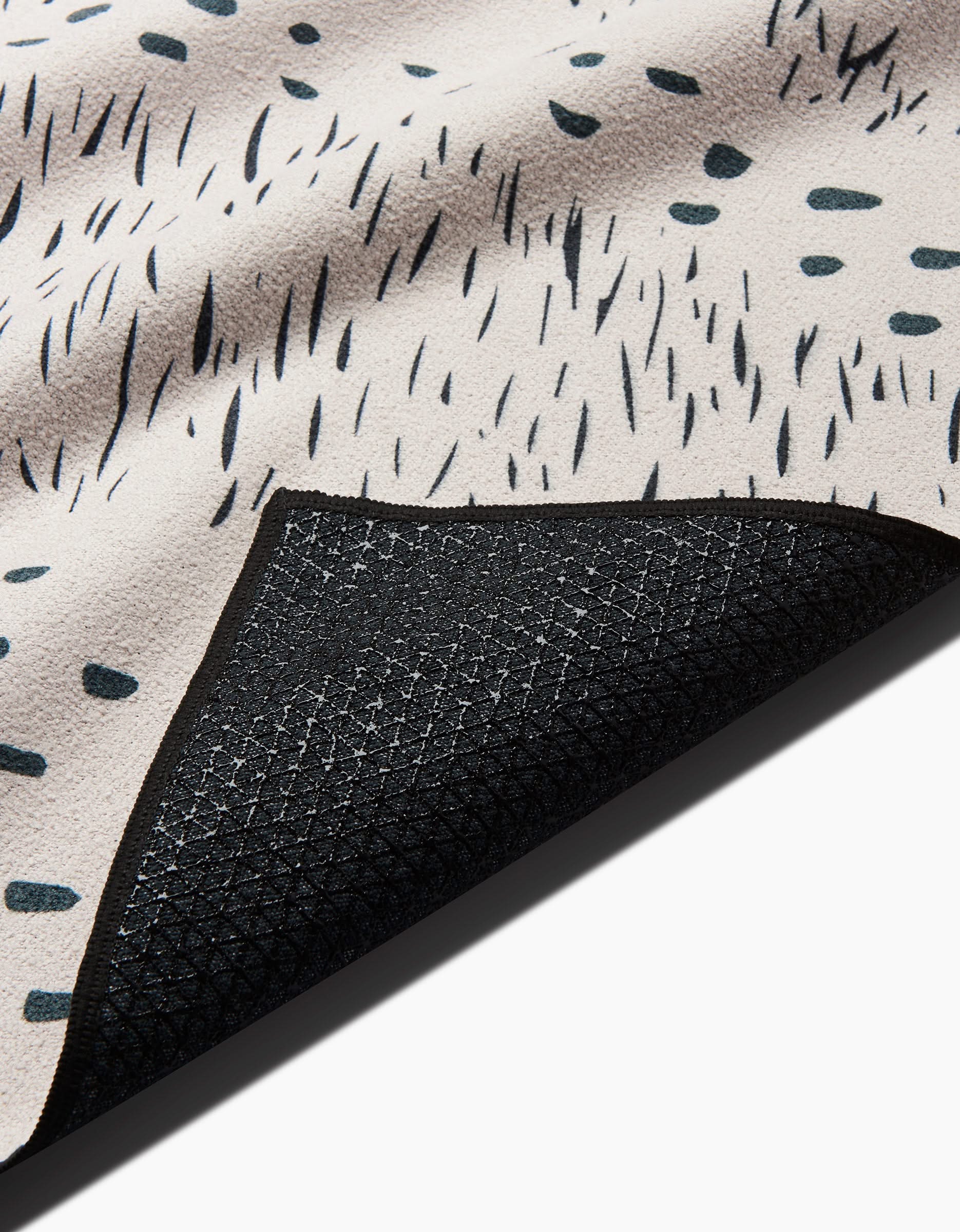 A close-up of the GEOMETRY Bend yoga towel shows its light beige surface with irregular black spots and a textured black non-slip backing.