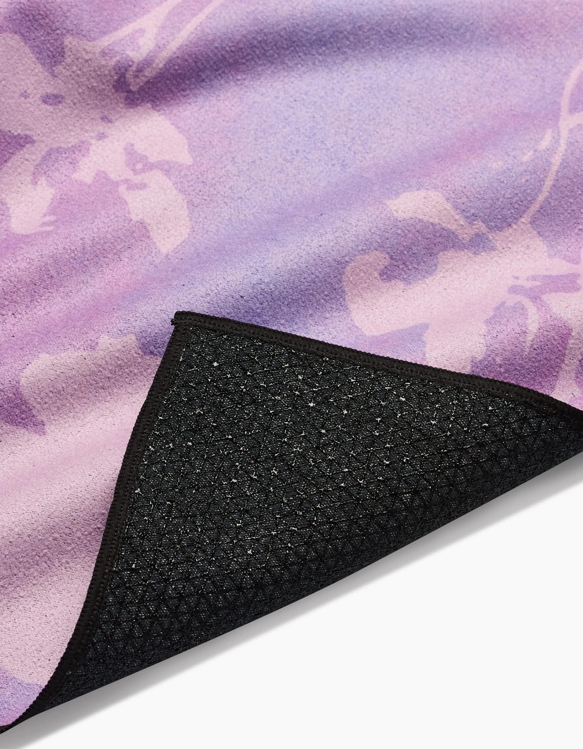 A close-up of the GEOMETRY Bloom Blend yoga mat, featuring a purple and pink floral pattern, crafted from recycled materials with a black textured non-slip backing; one corner is slightly lifted to show the underside.